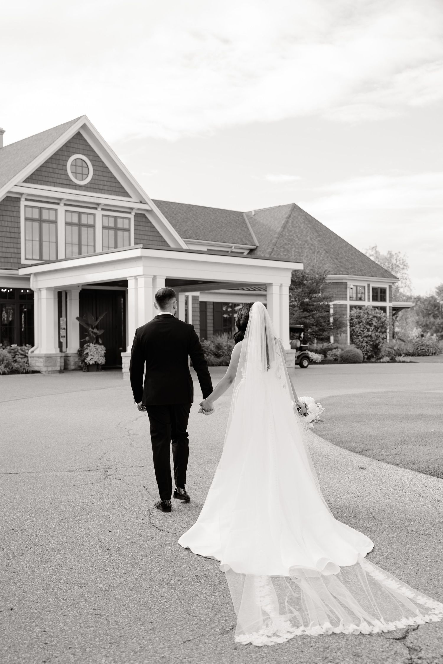 A wedding couple walks away from a gray shingle-style house with a long flowing white veil trailing behind them.