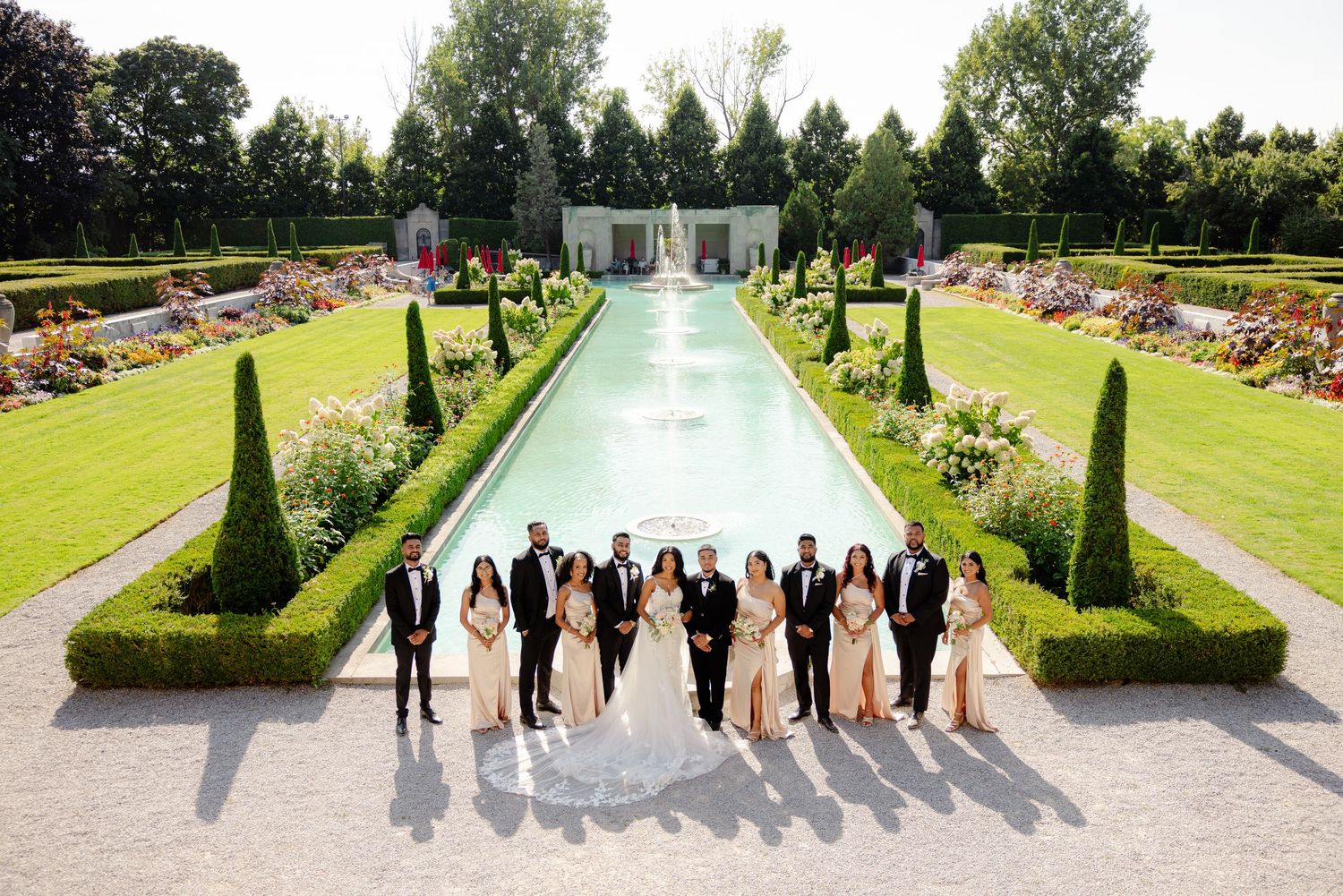 Wedding party lined up along a formal garden pool with cypress trees and colorful flower beds.