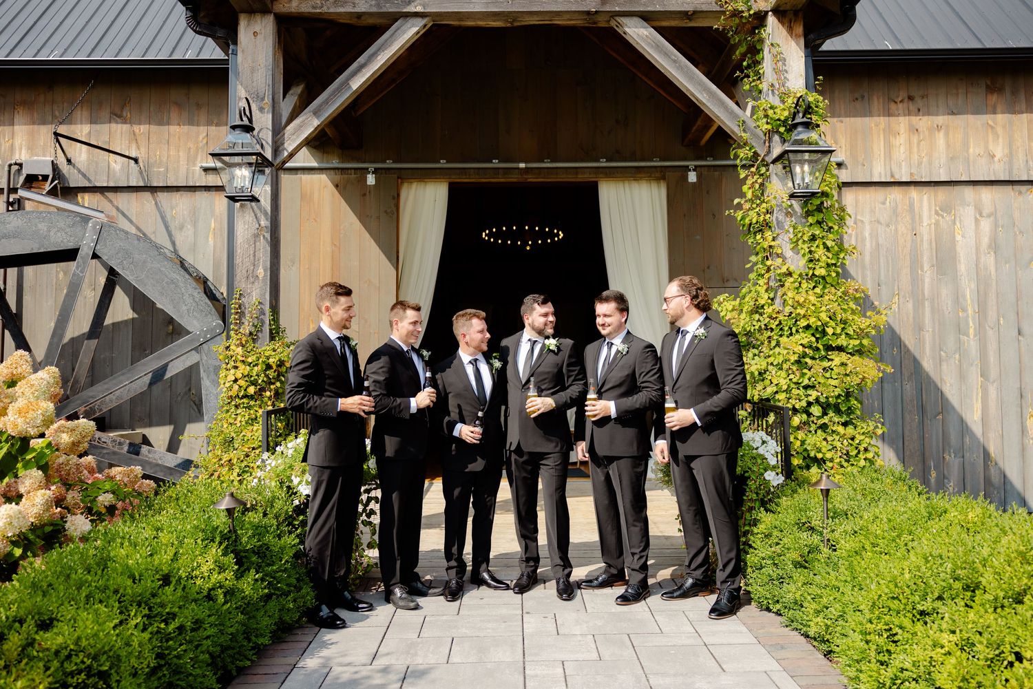 Wedding party shares drinks and laughter outside rustic barn entrance with green foliage.