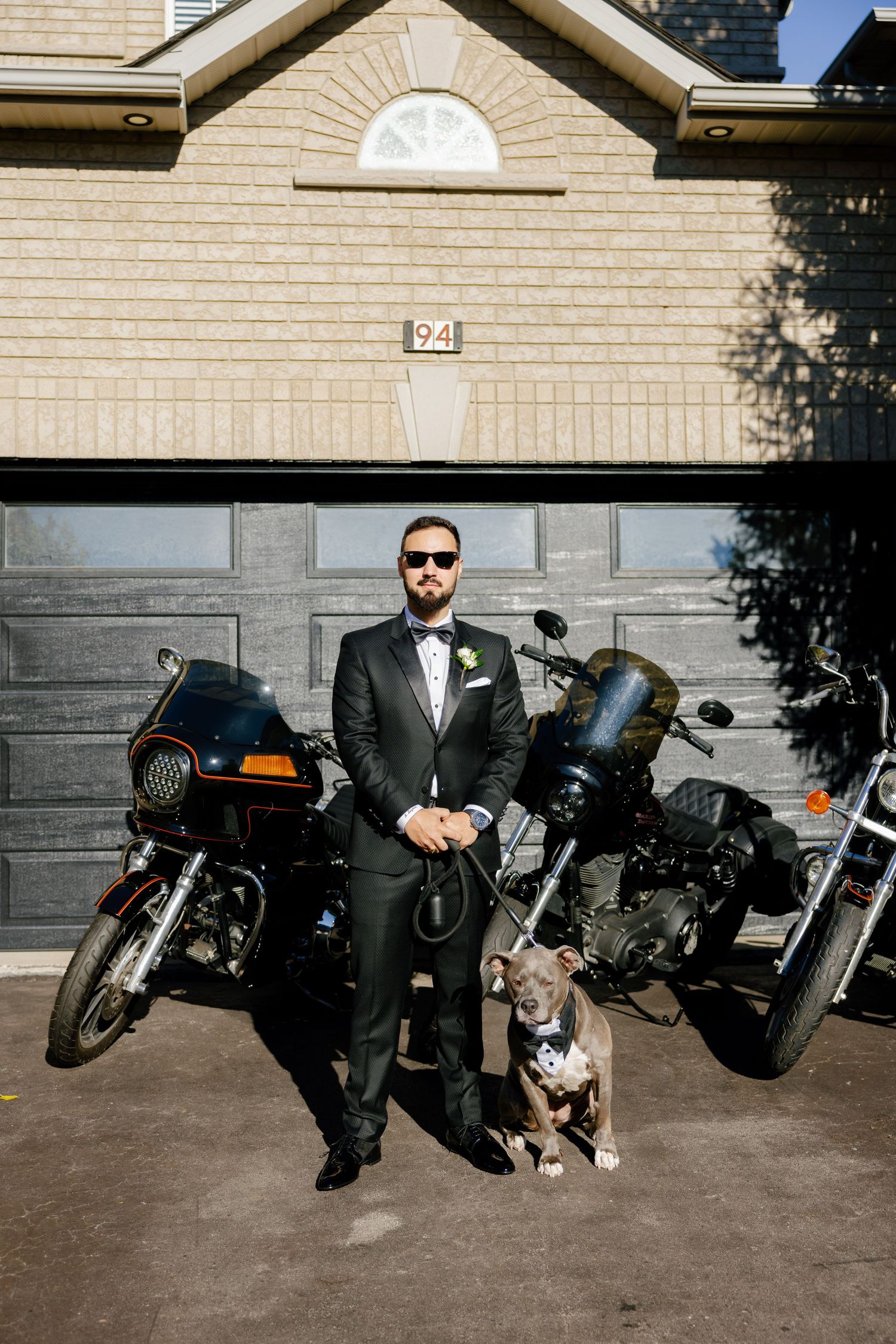A dapper groom in a black suit poses with his dog and motorcycles in front of a garage door.