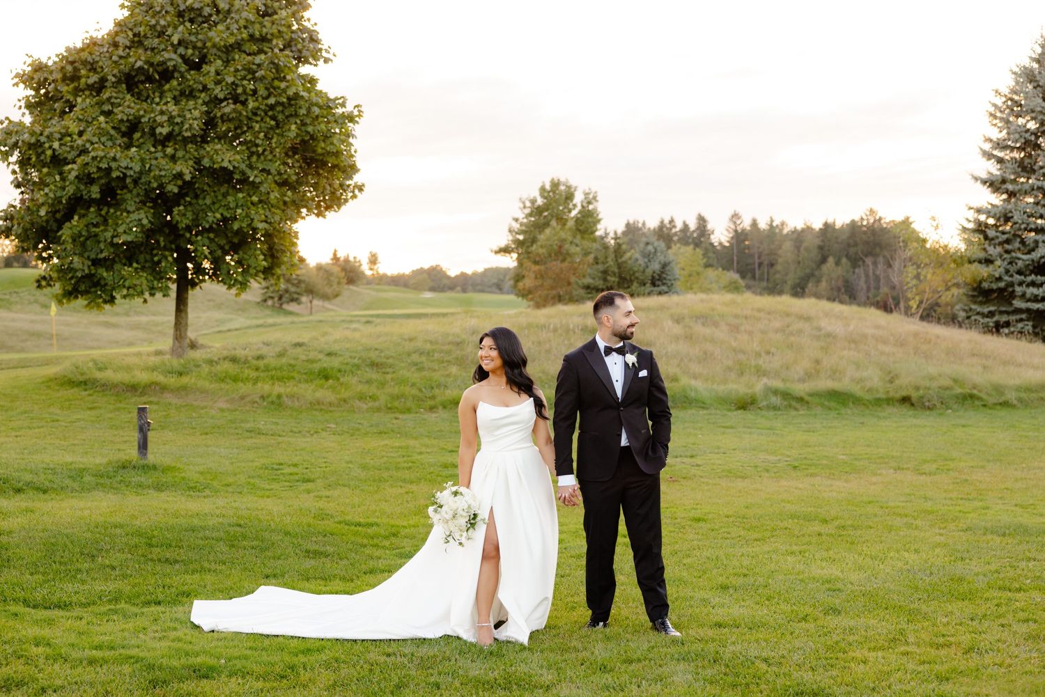 Couple standing on hillside with elegant white gown and tall tree backdrop.