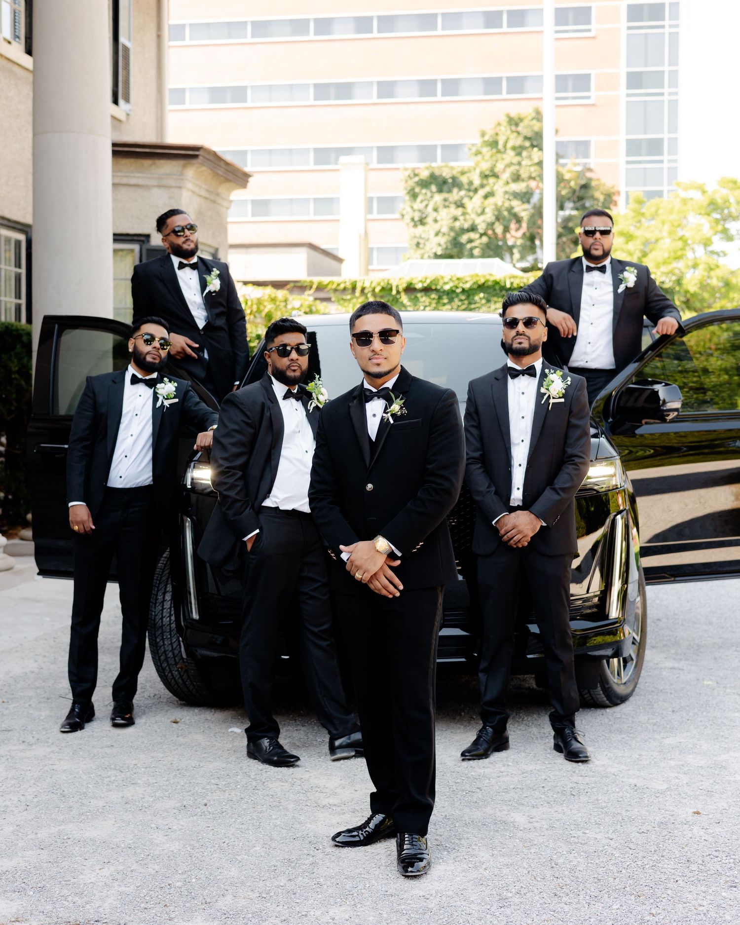 Wedding party in black tuxedos poses with sunglasses near a luxury vehicle outside a classic building.