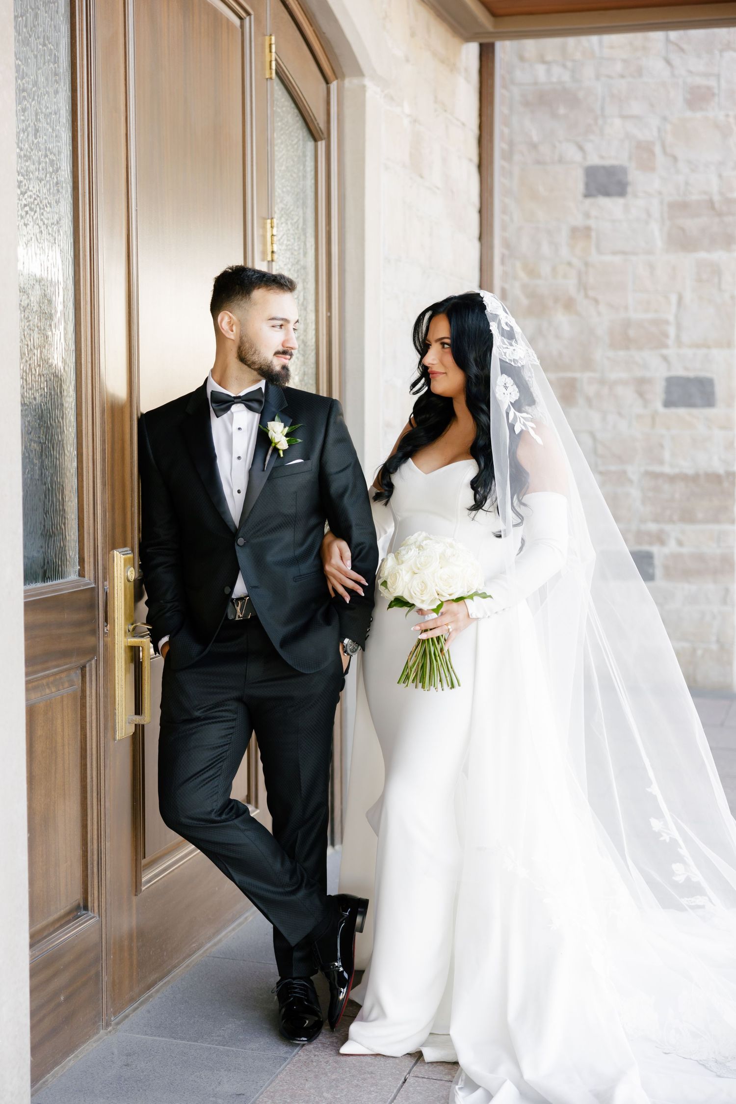 Wedding couple in classic black tuxedo and white gown pose by wooden doors.