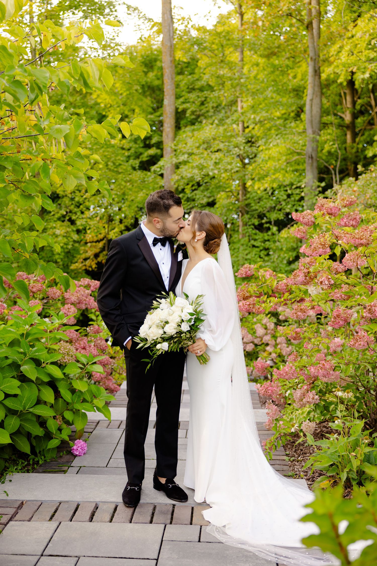A romantic wedding couple embraces on a garden path surrounded by pink flowers and lush greenery.