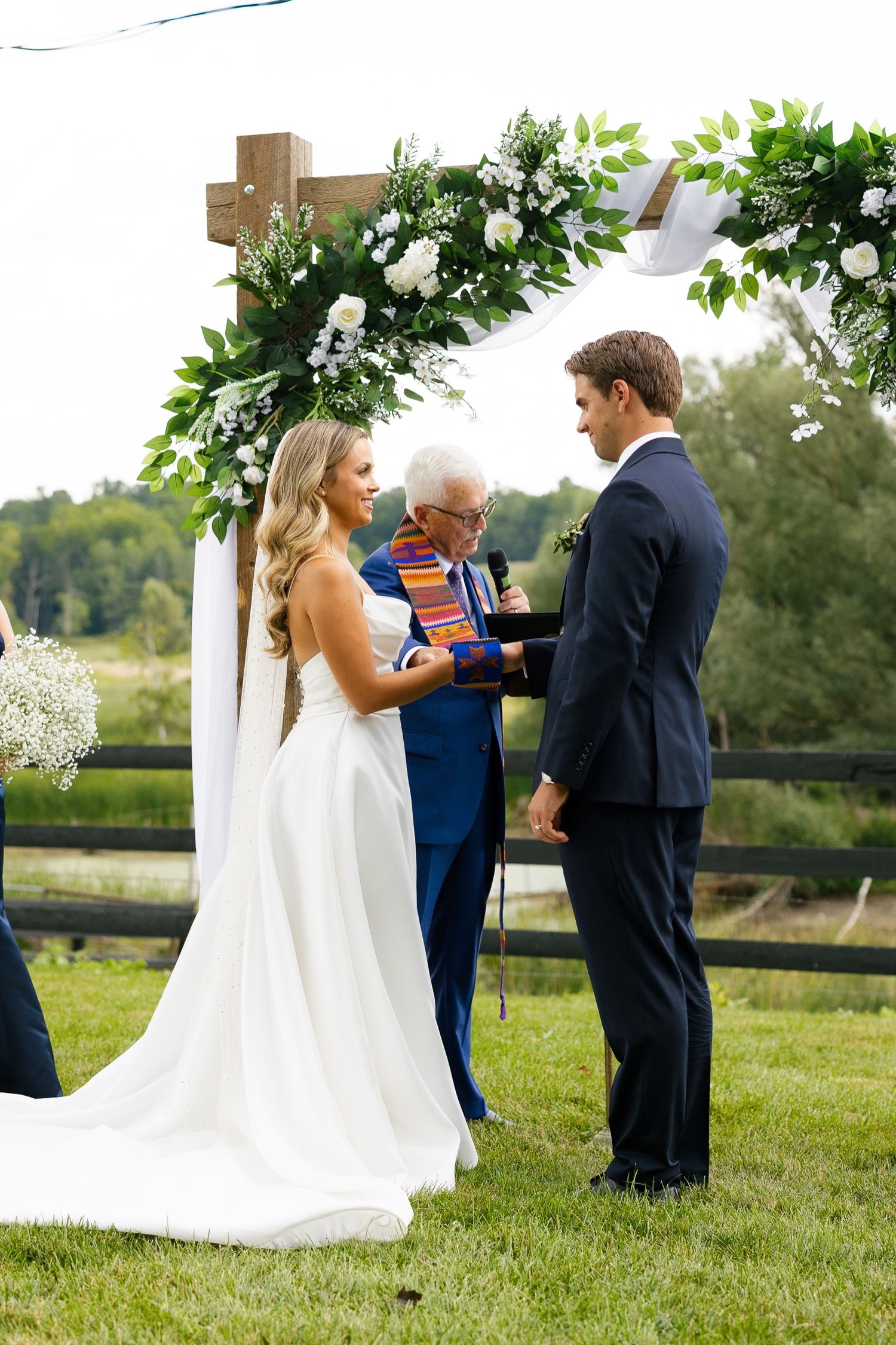 A wedding ceremony unfolds beneath a rustic wooden arch decorated with white roses and foliage.