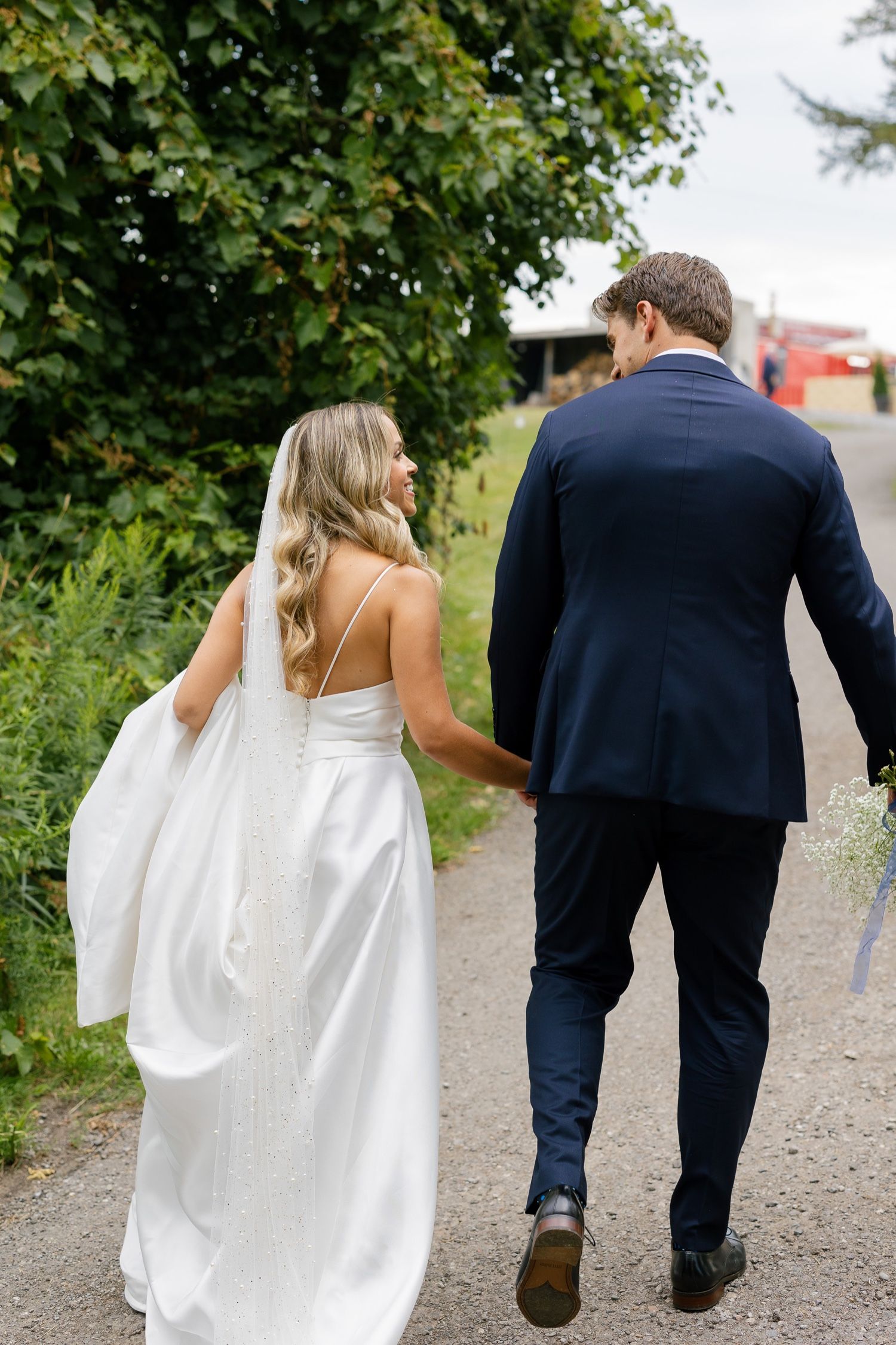 A bride and groom walk hand in hand down a country path, surrounded by greenery.