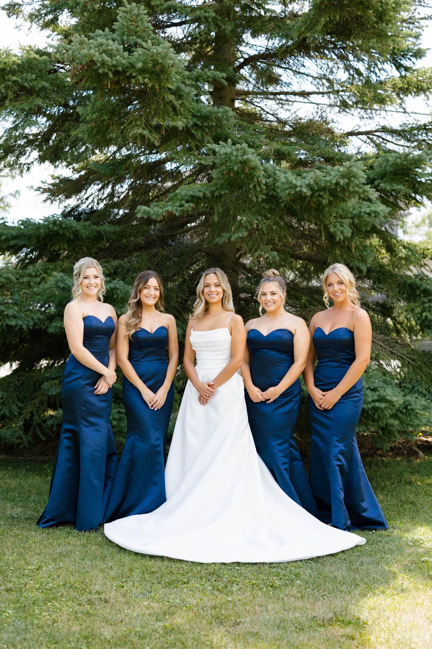 Bridal party in navy blue dresses poses formally against a large evergreen tree.