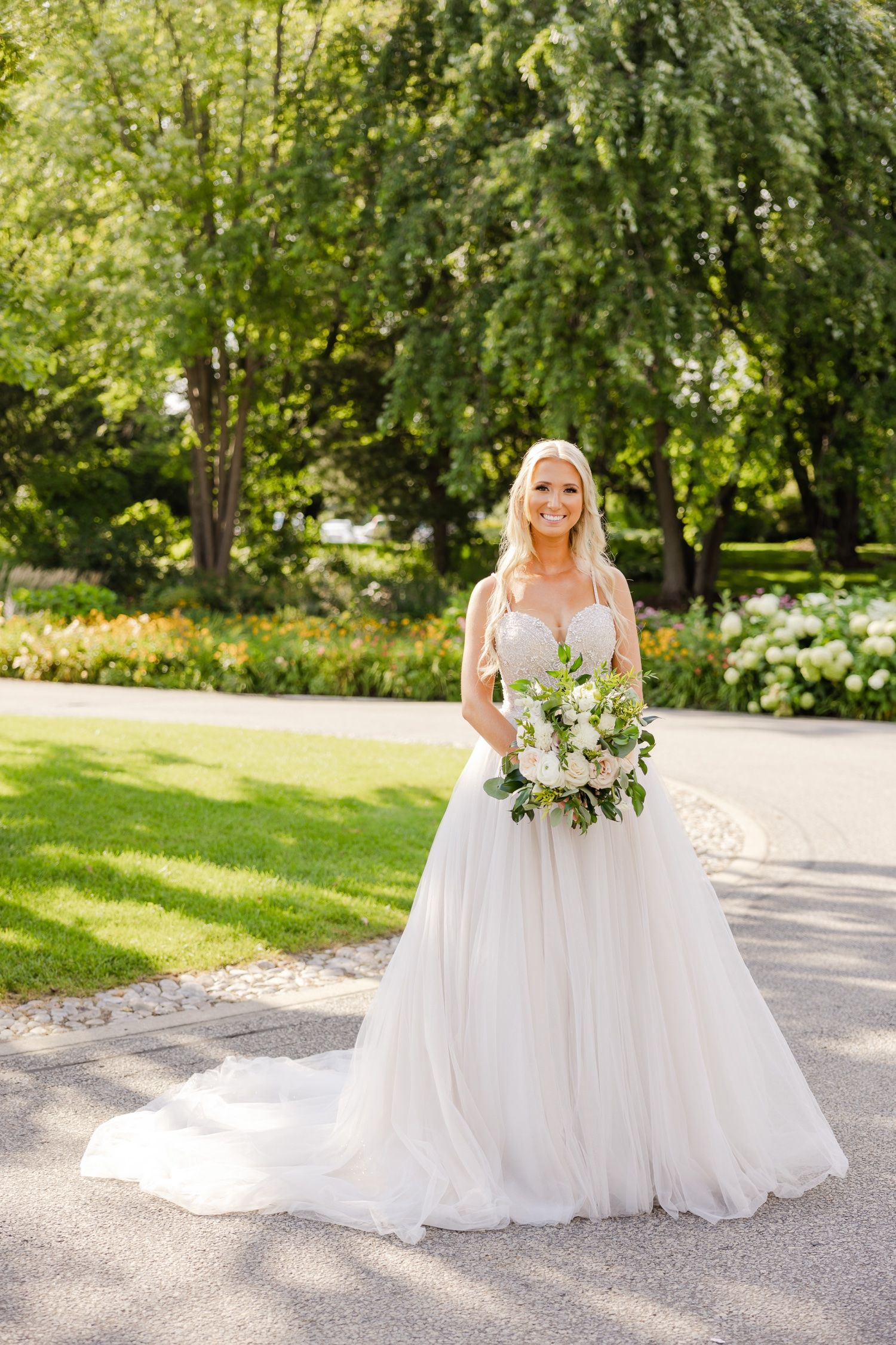 A bride in a flowing white ballgown poses in a lush garden setting holding a white and green bouquet.