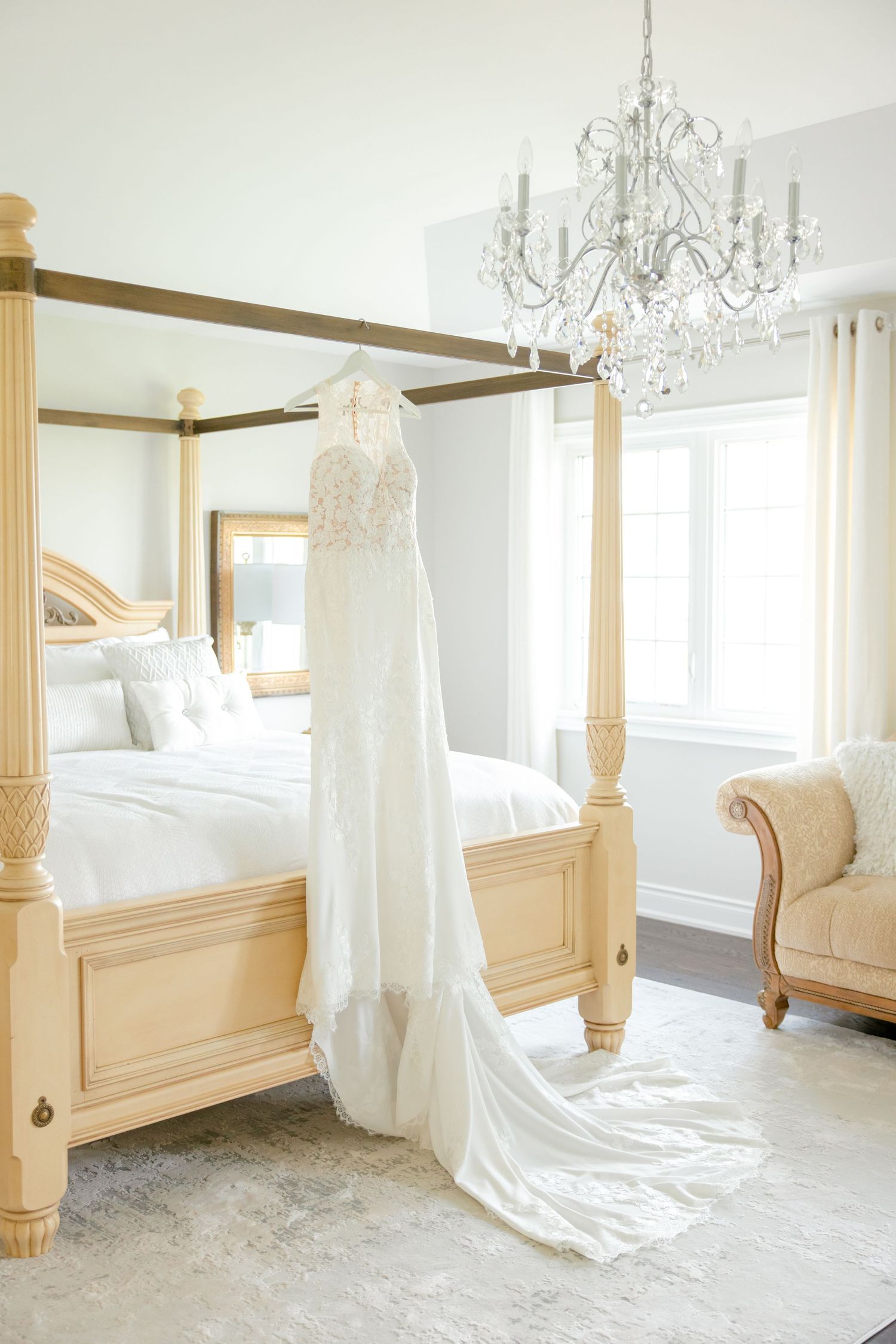 Elegant lace wedding dress hanging from four-poster bed under crystal chandelier in bright white room.