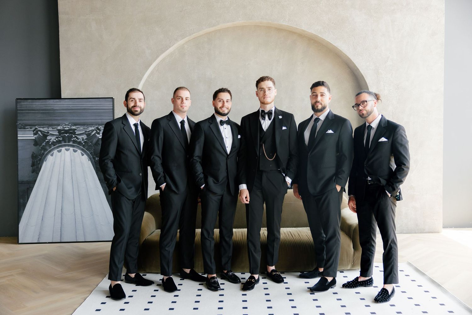 Groomsmen in dark suits and ties pose together against a light wall with an arched alcove.