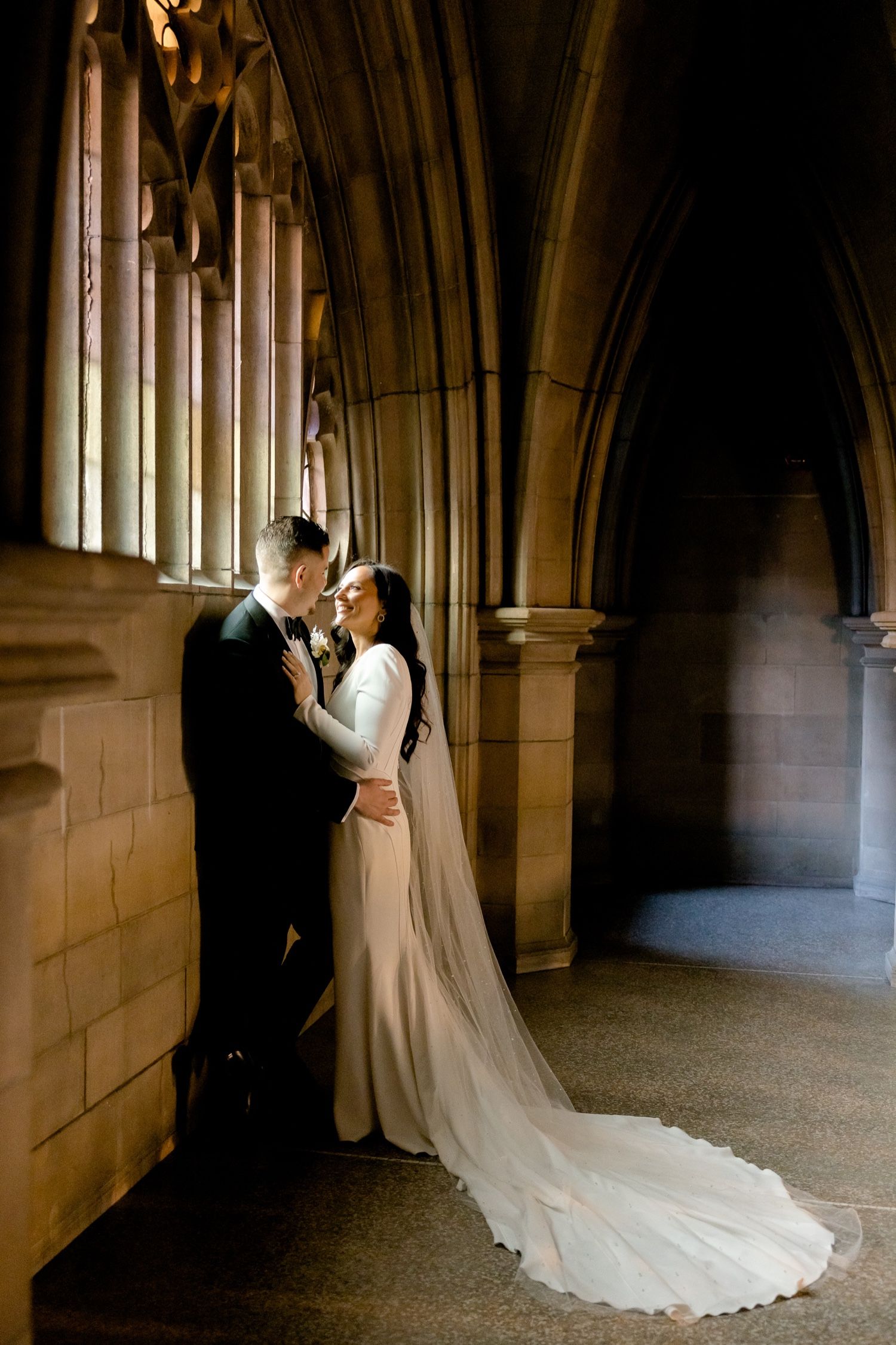 Elegant wedding portrait against gothic stone columns with flowing white train and dramatic lighting.