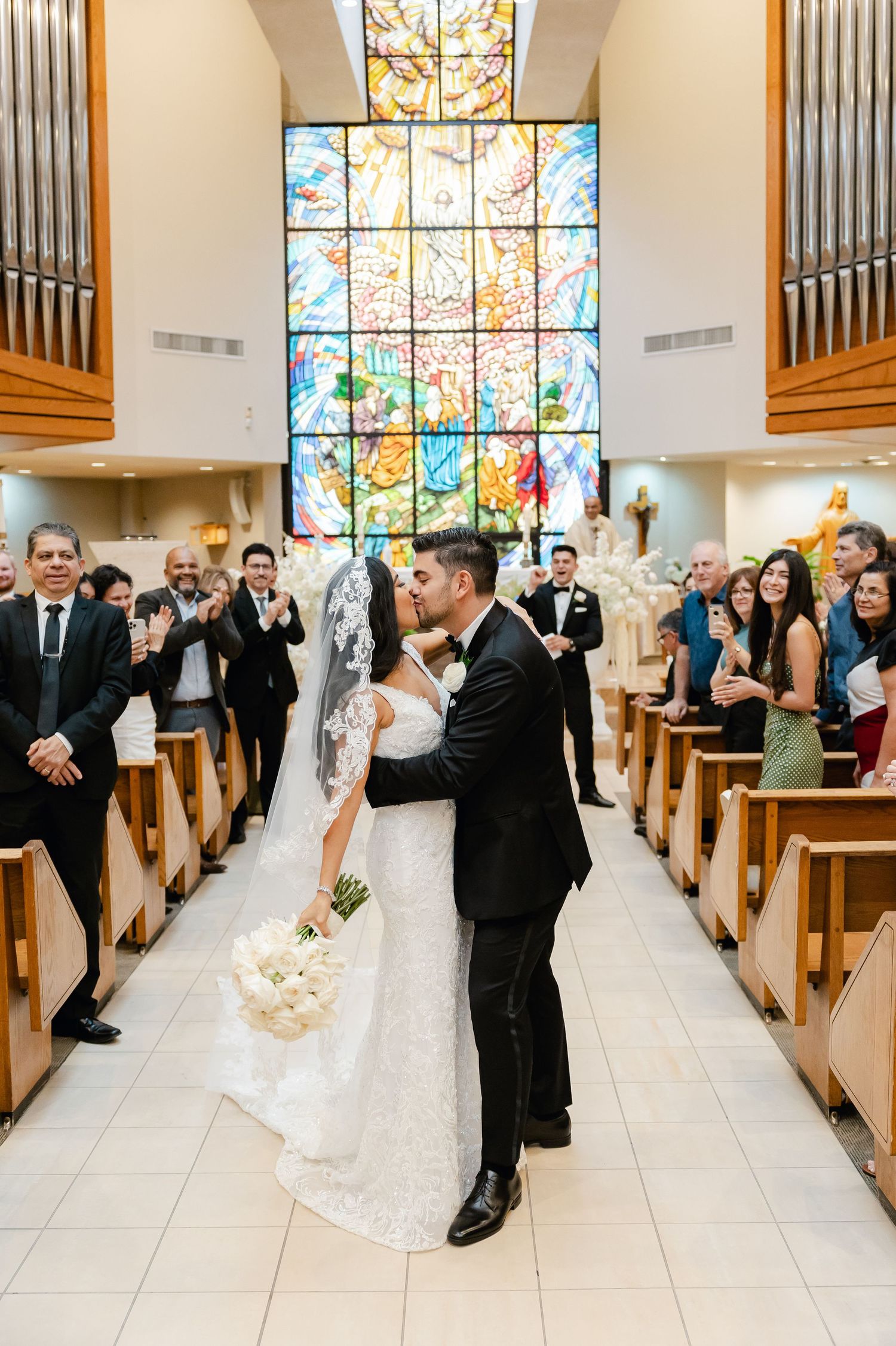 Wedding couple sharing romantic kiss in church aisle as guests applaud.