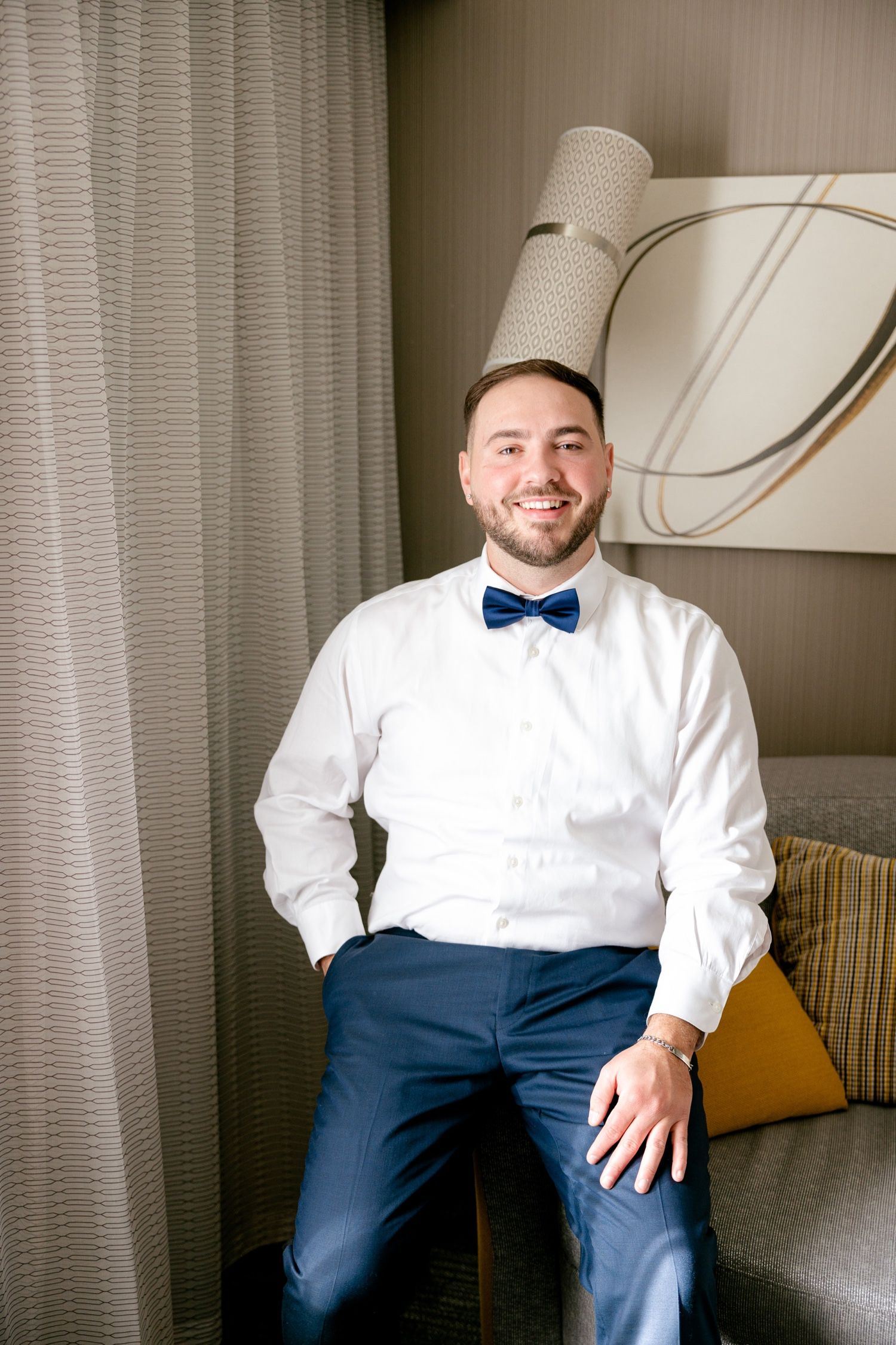 A person in formal attire wearing a white dress shirt and navy blue bow tie sits on a gray couch.
