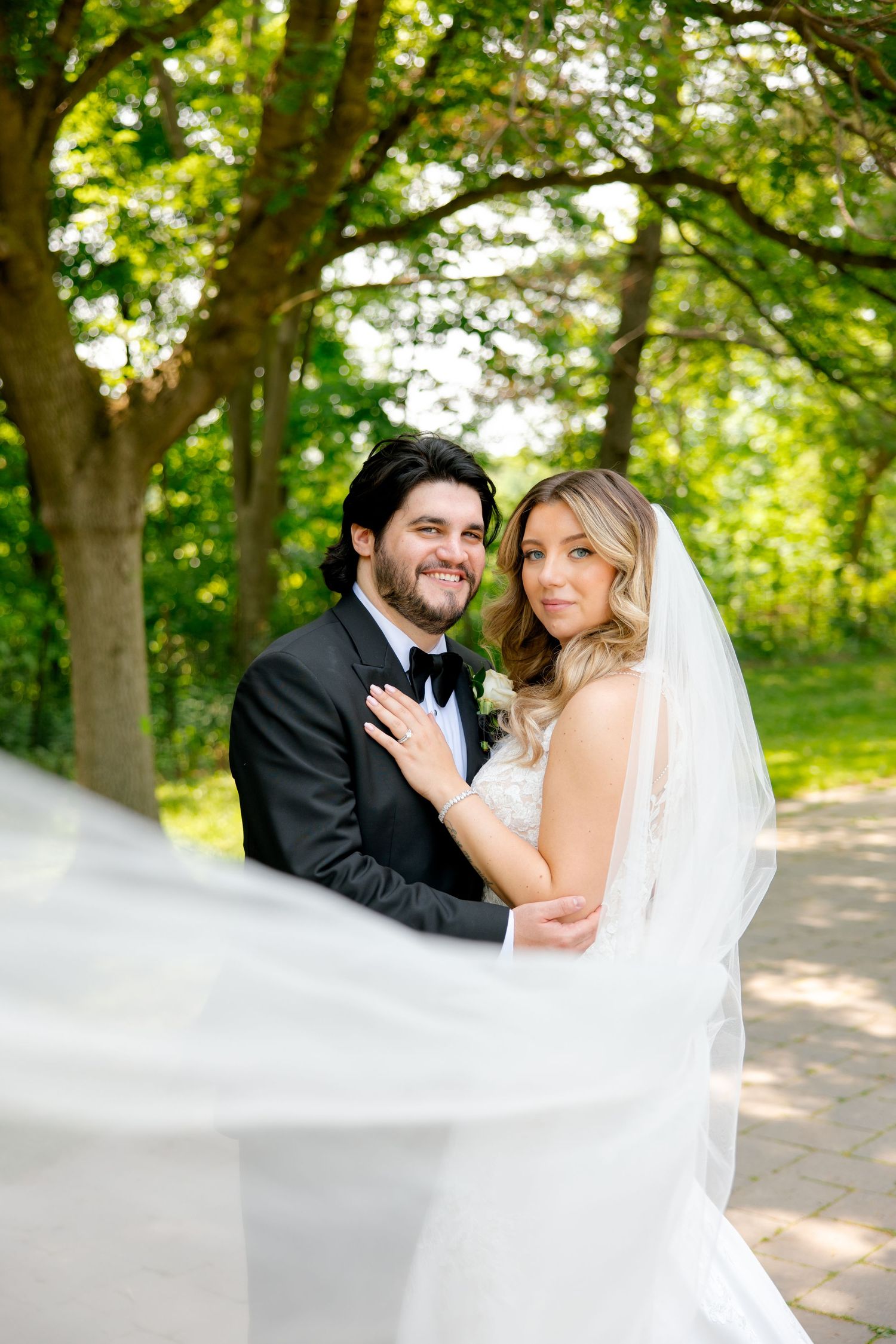 A joyful wedding portrait with the bride's veil floating in the breeze against green trees.