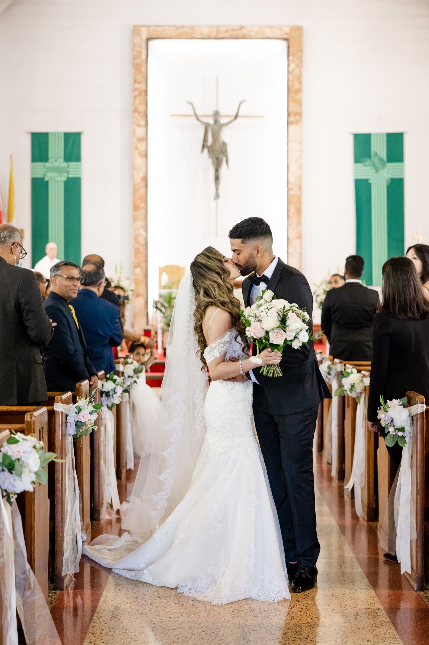 A romantic wedding kiss in a church aisle decorated with white flowers and green banners.