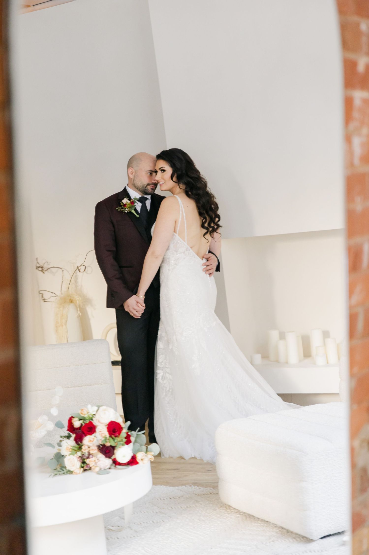 An elegant wedding couple poses in a modern white room with exposed brick walls.