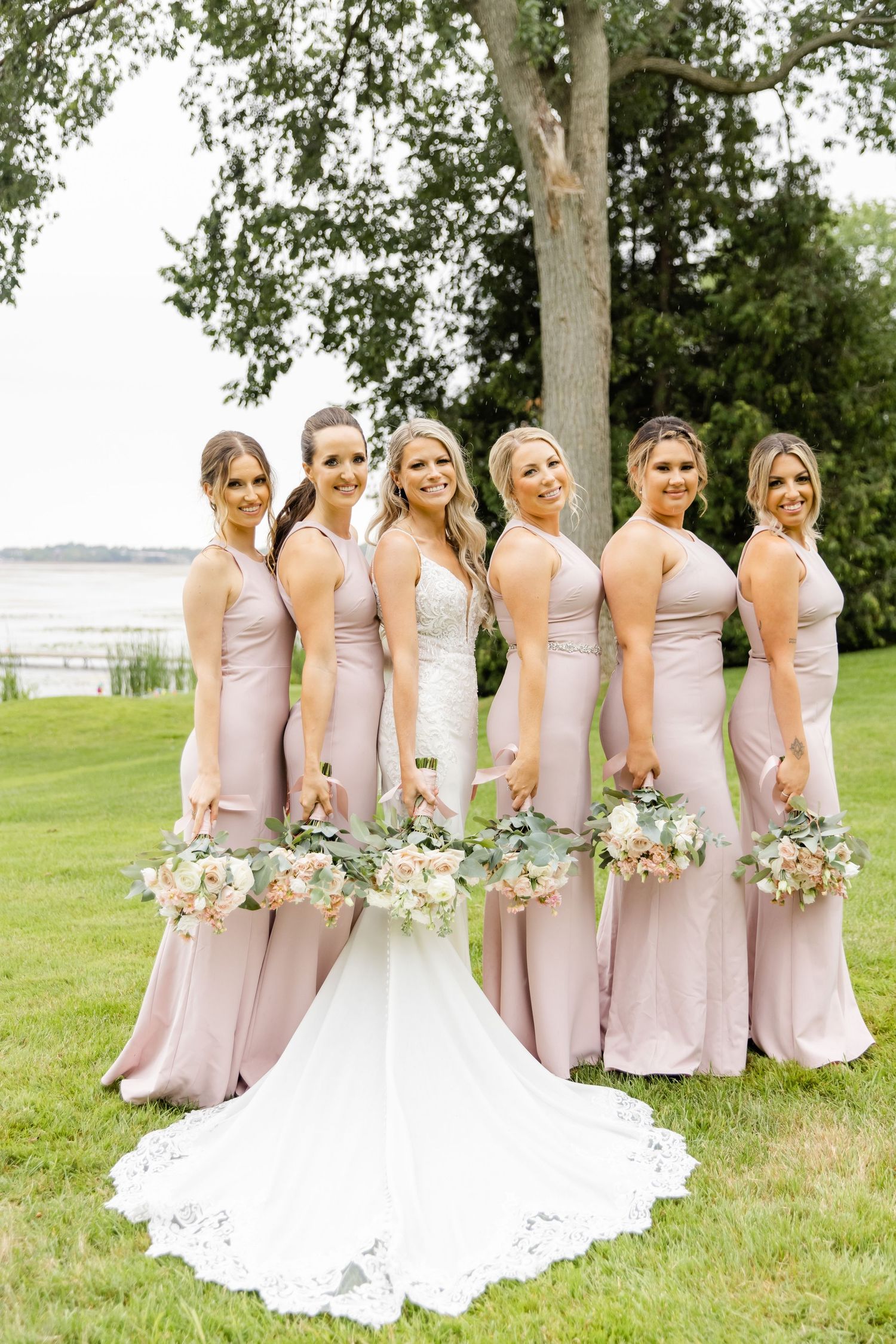 Wedding party in blush gowns and white dress pose elegantly on waterfront lawn.