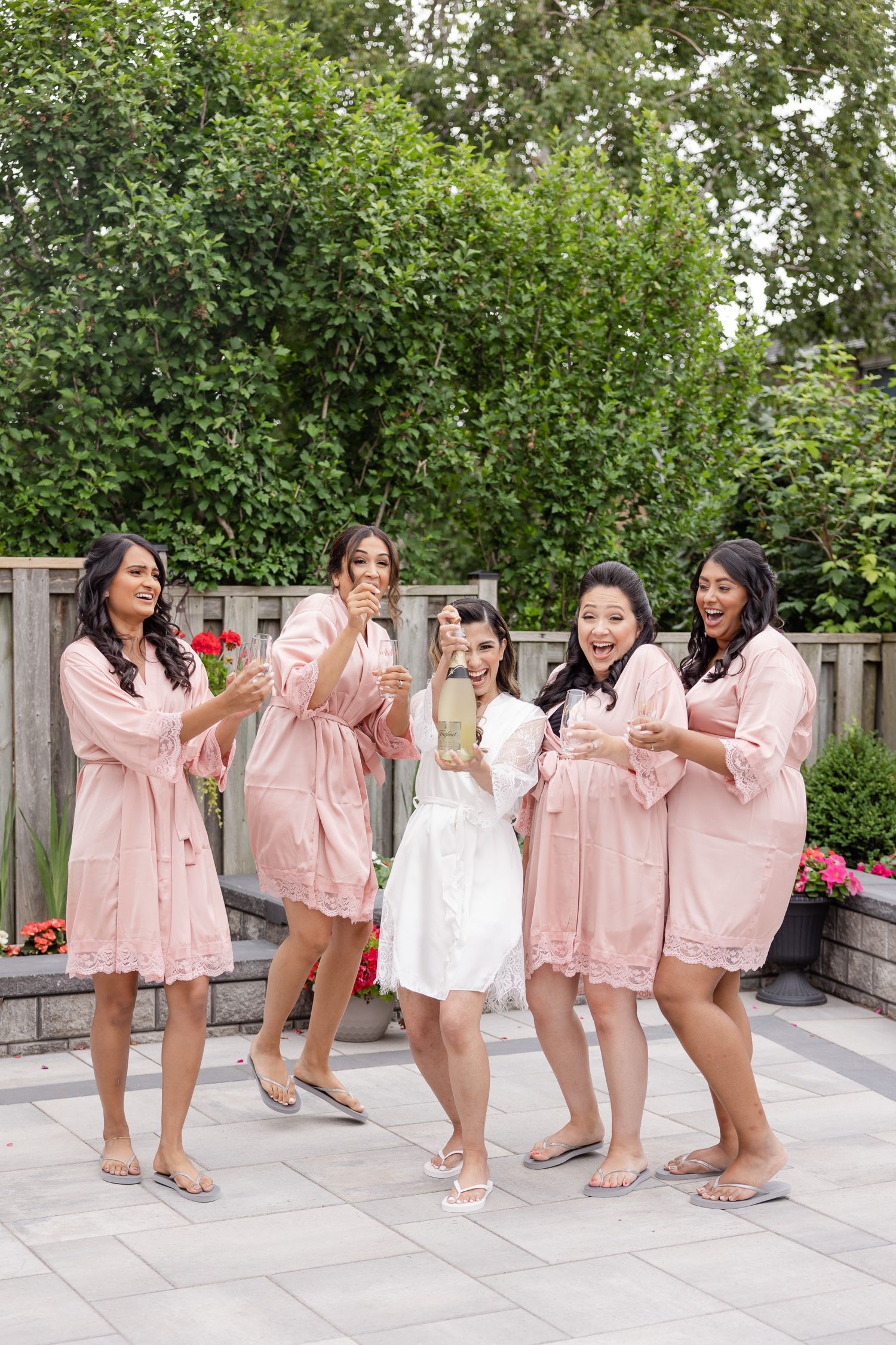 Wedding party in pink robes laughing and celebrating with champagne on outdoor patio.