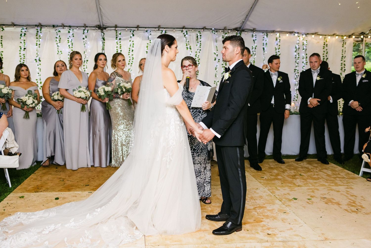 Wedding ceremony in progress under white tent with string lights and floral decorations.