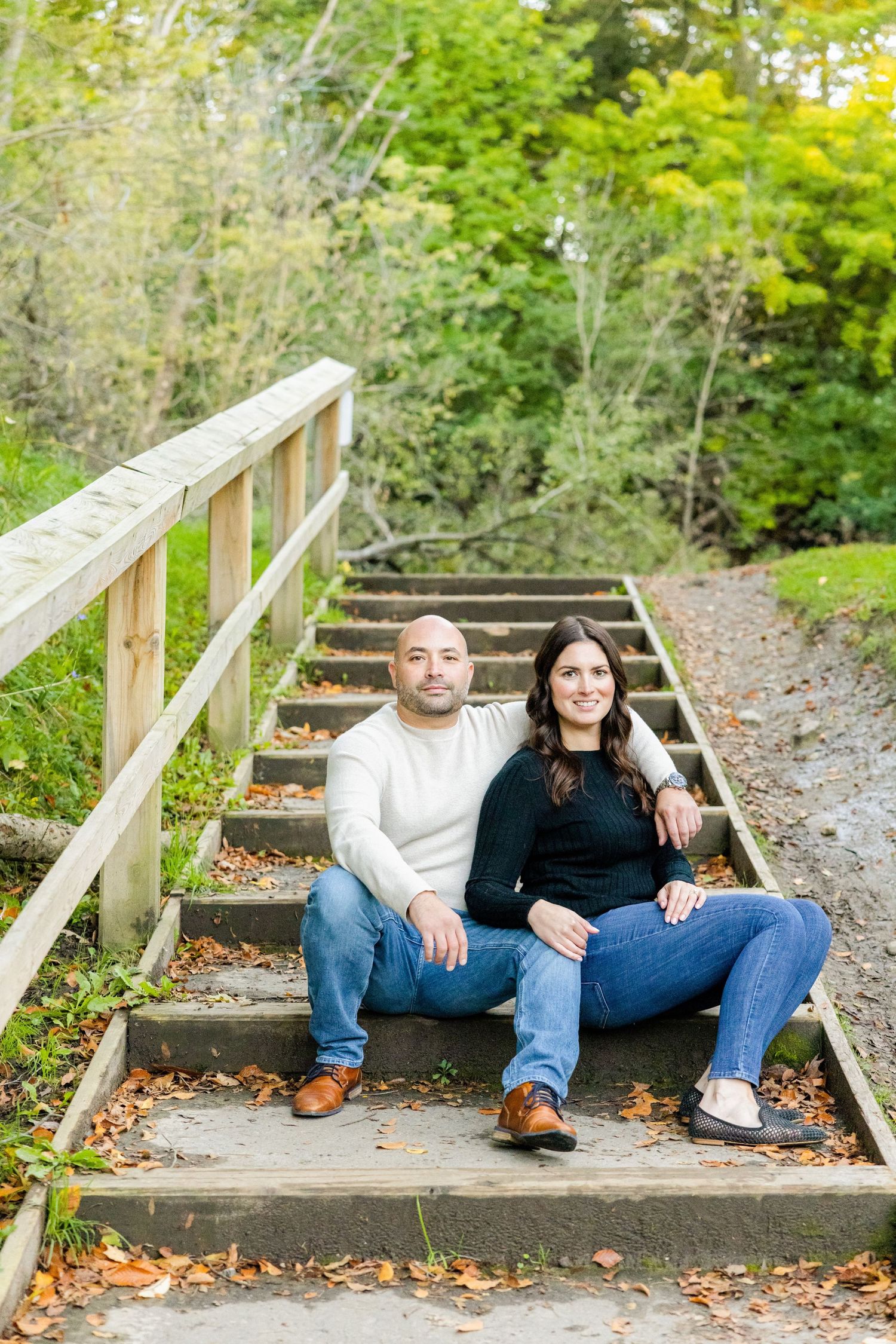 A couple relaxes on outdoor wooden steps surrounded by lush greenery and fallen leaves.