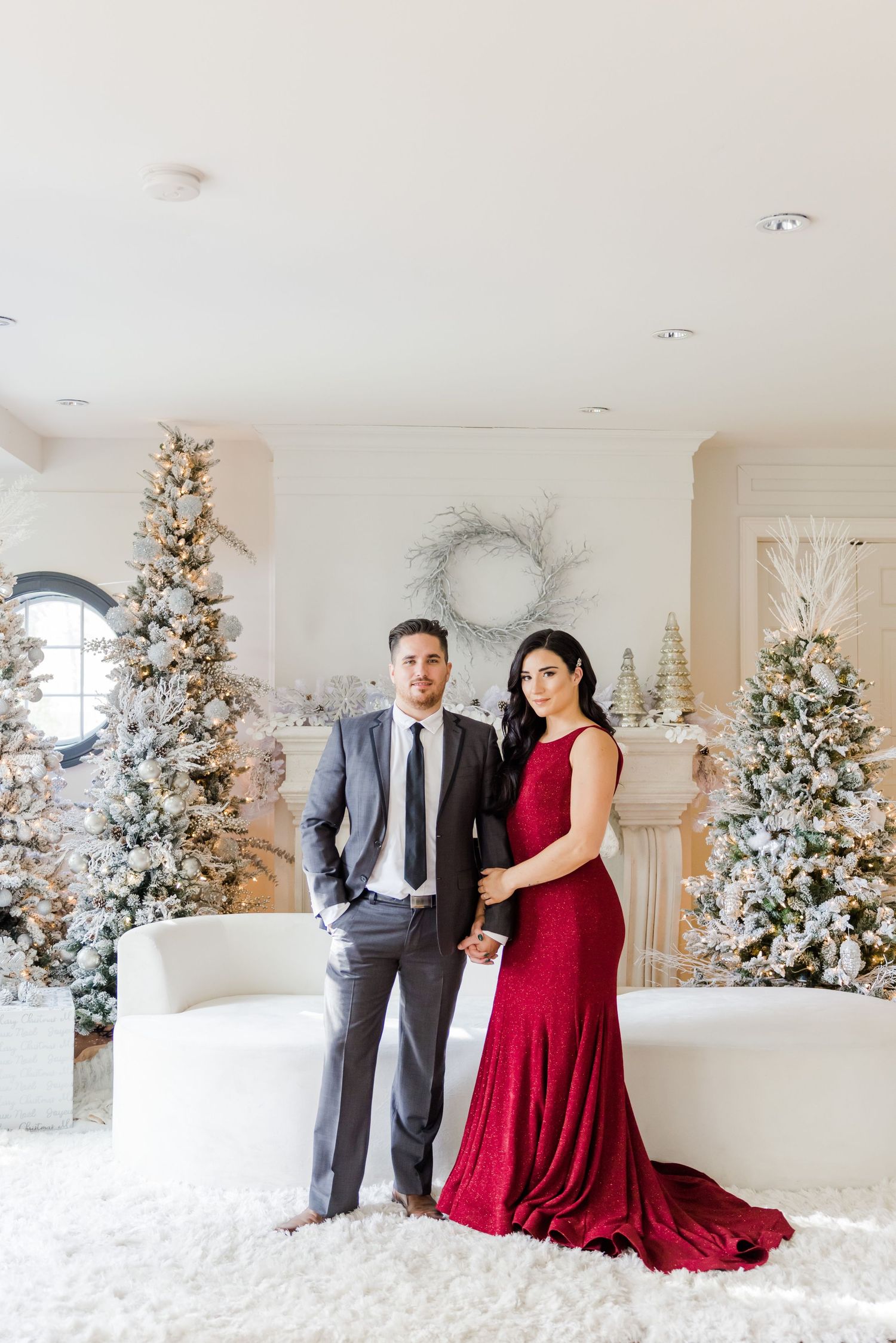 Full-length portrait of couple in formal wear standing among decorated Christmas trees in bright white room.