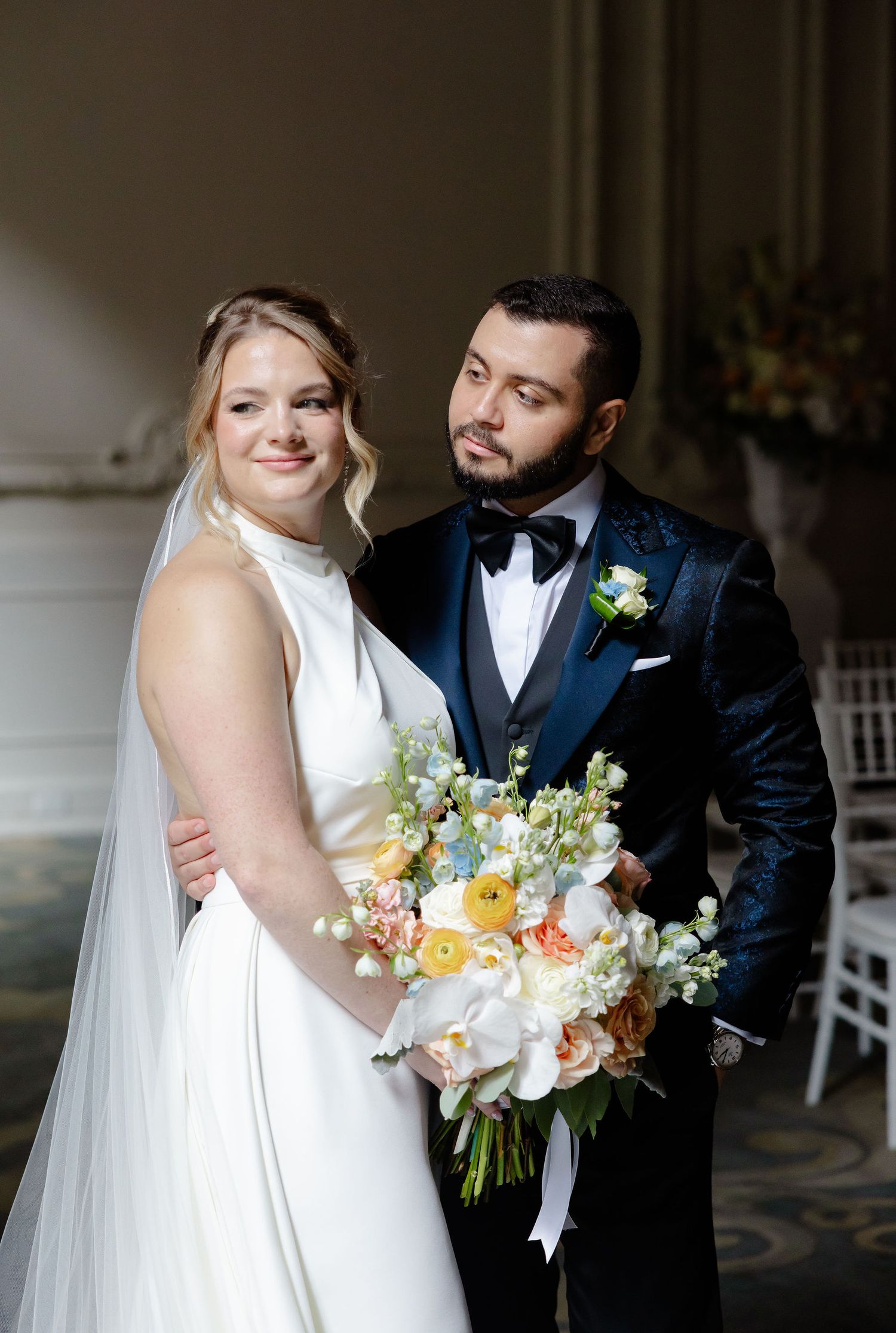 Bride in white gown holds peach and white bouquet while sharing intimate moment with groom.