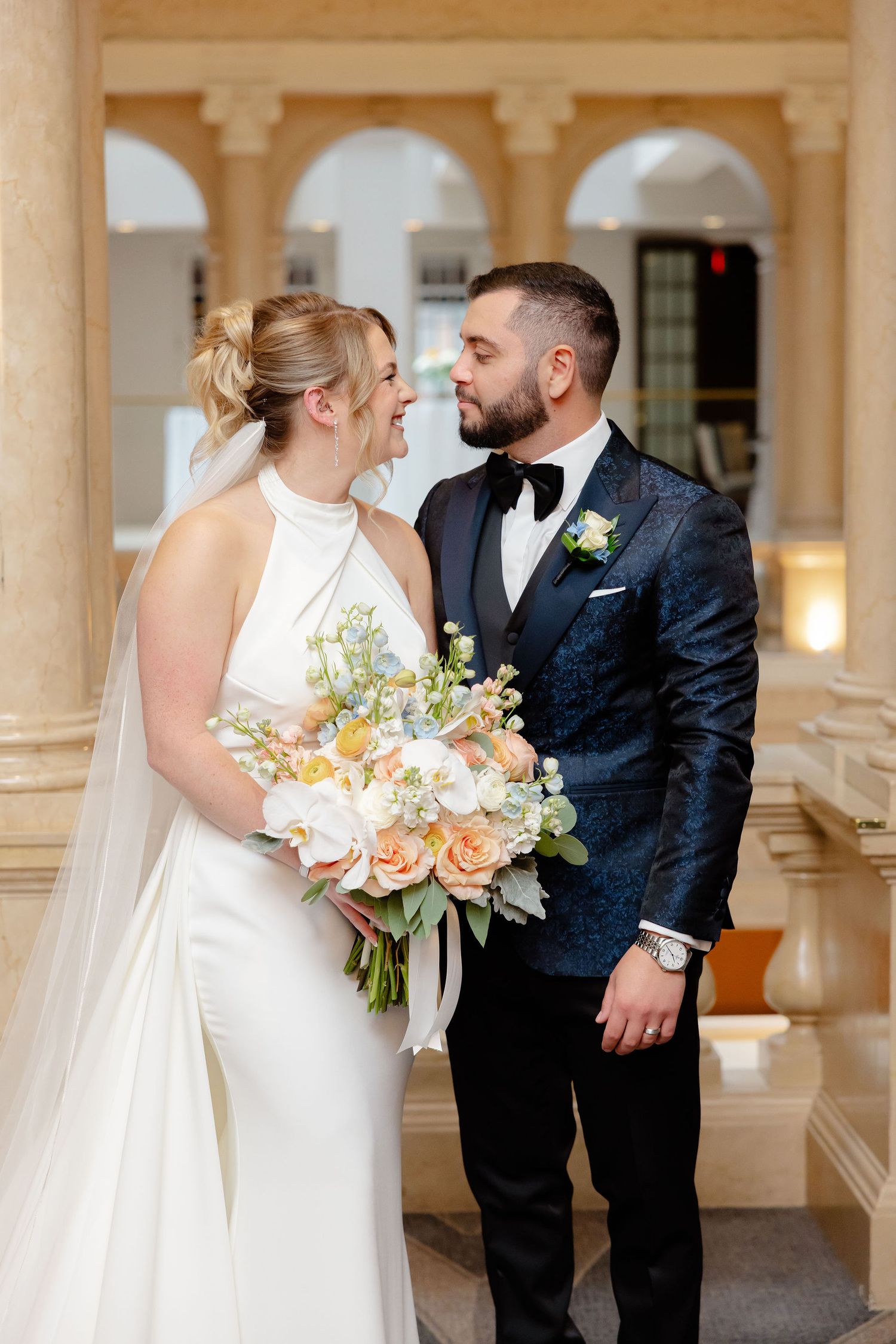 Bride holding peach and white bouquet shares intimate moment with groom in front of classical columns.