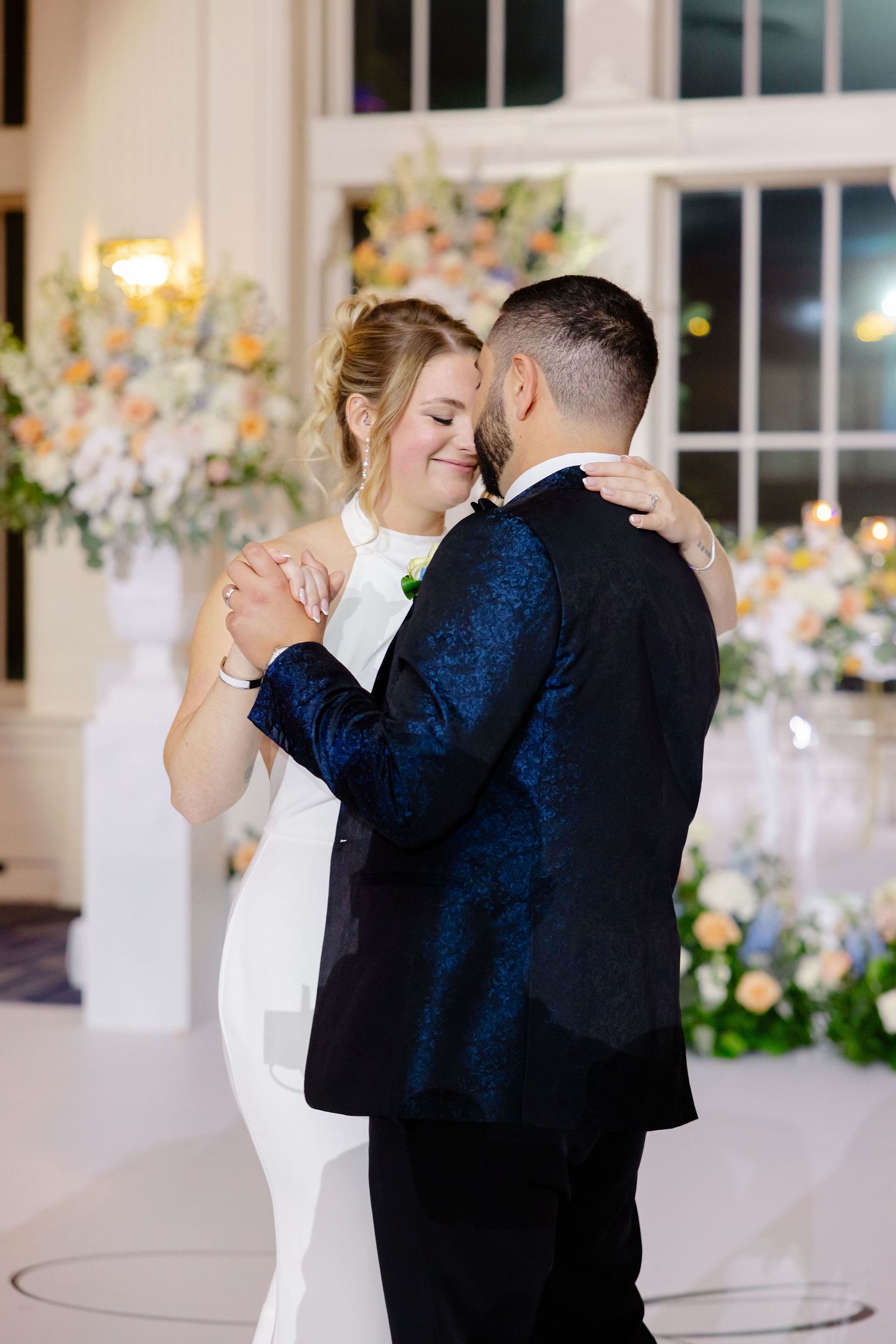 Couple sharing an intimate dance at an elegant wedding reception with floral decorations and large windows.