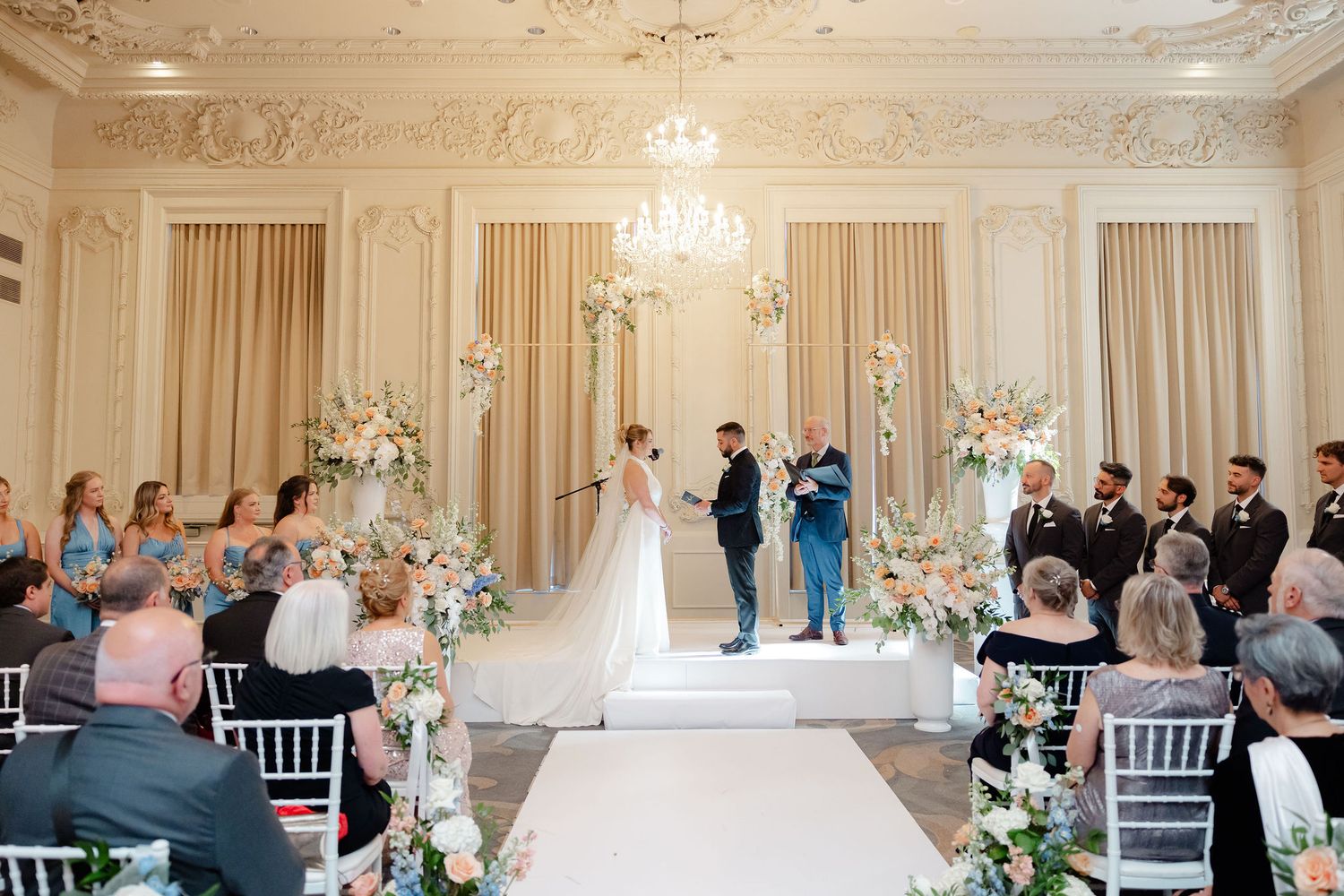 Wedding ceremony in progress in an ornate ballroom with white floral decorations and crystal chandelier.
