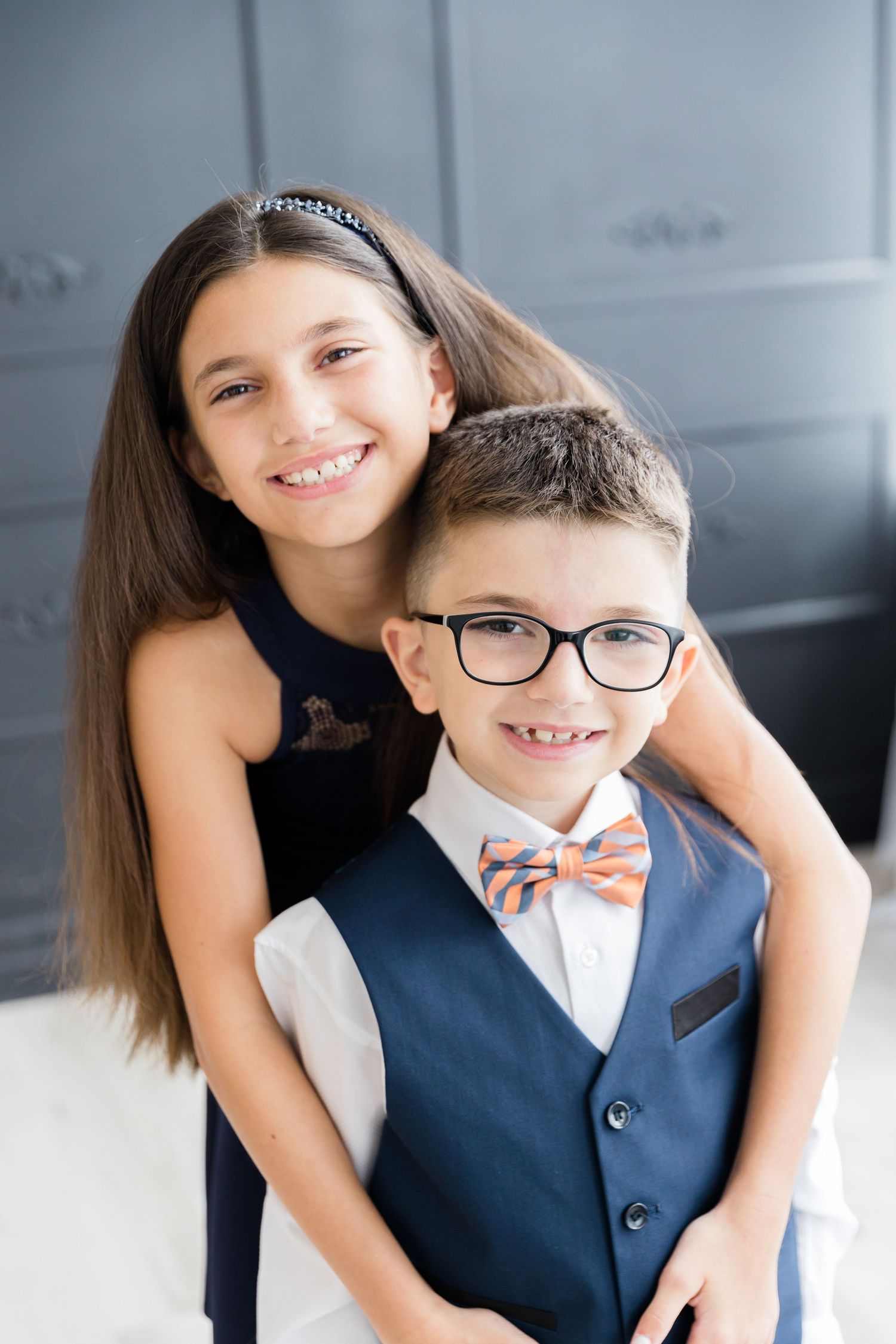 Close-up portrait of siblings smiling in navy formal wear against a gray paneled background.