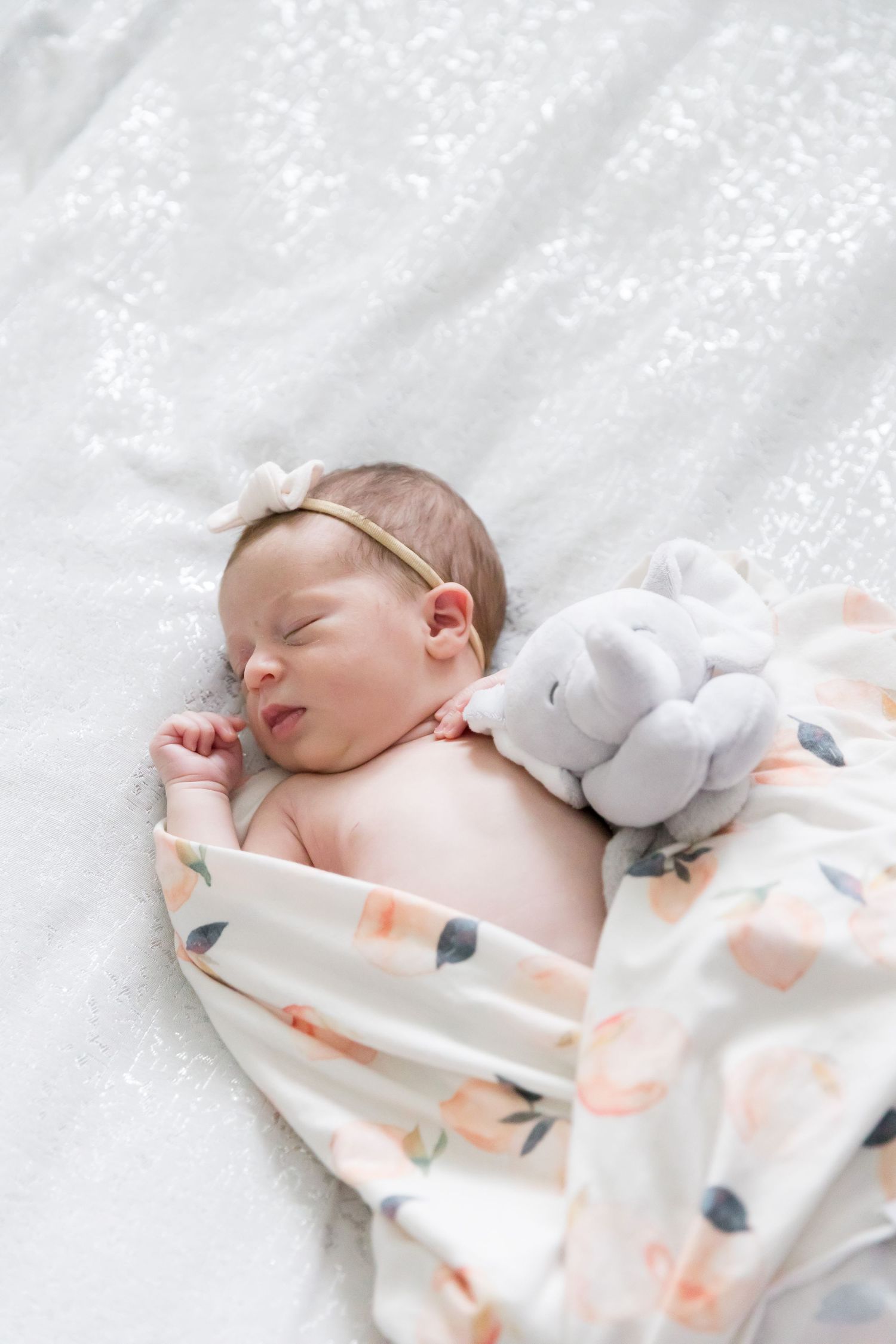 Peaceful sleeping newborn on white bedding with a small plush toy and floral swaddle.