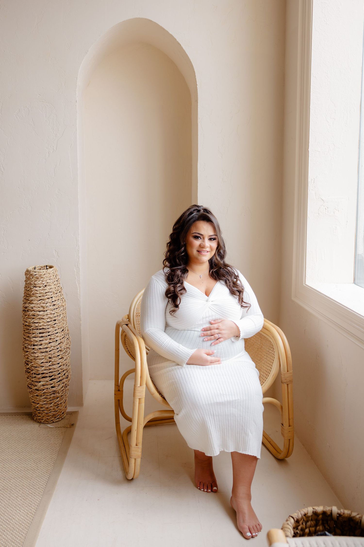A person in a white knit dress sits in a rattan chair by a window with a decorative vase nearby.
