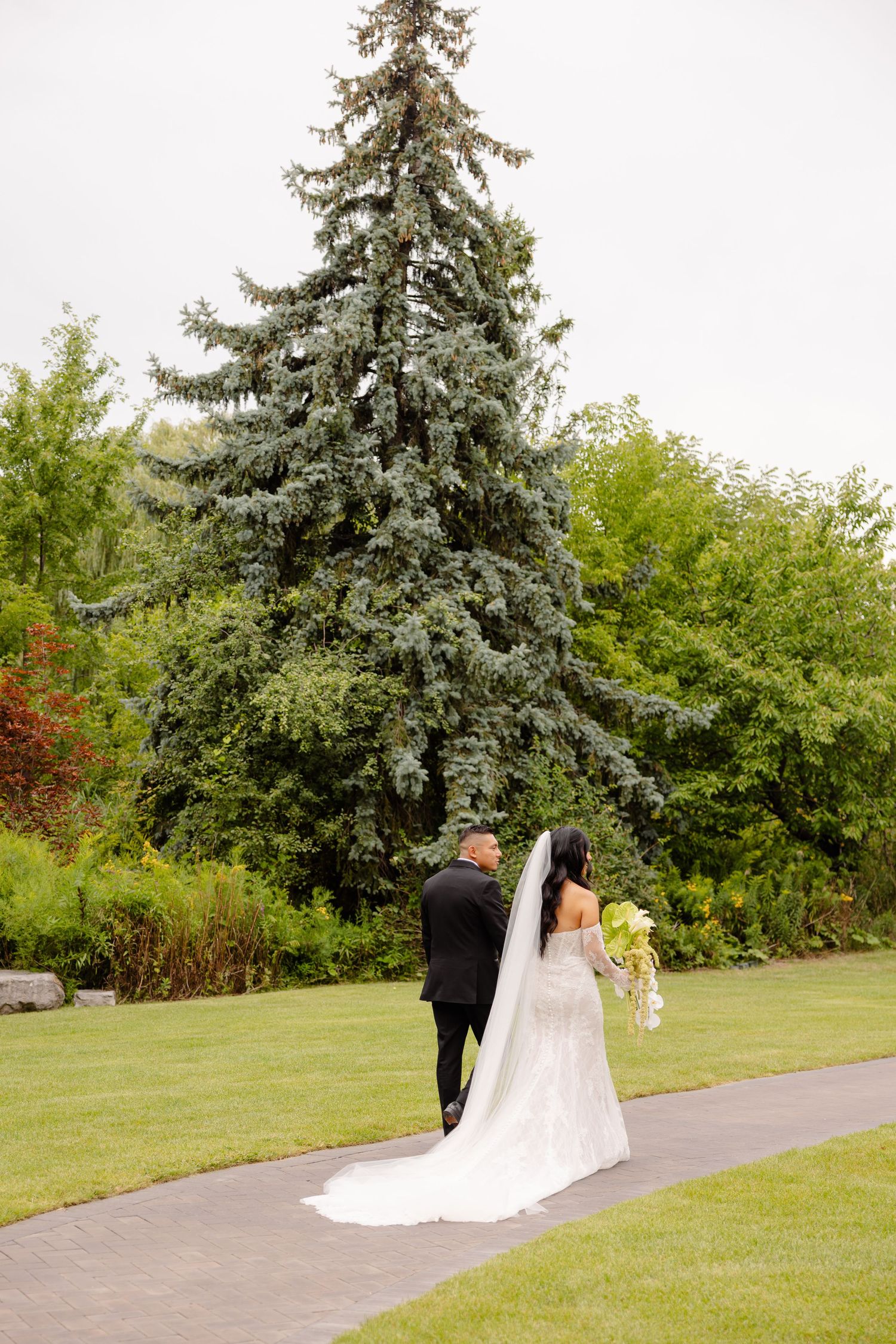 A couple walks together on a paved garden path surrounded by evergreen trees and lush greenery on a summer day.