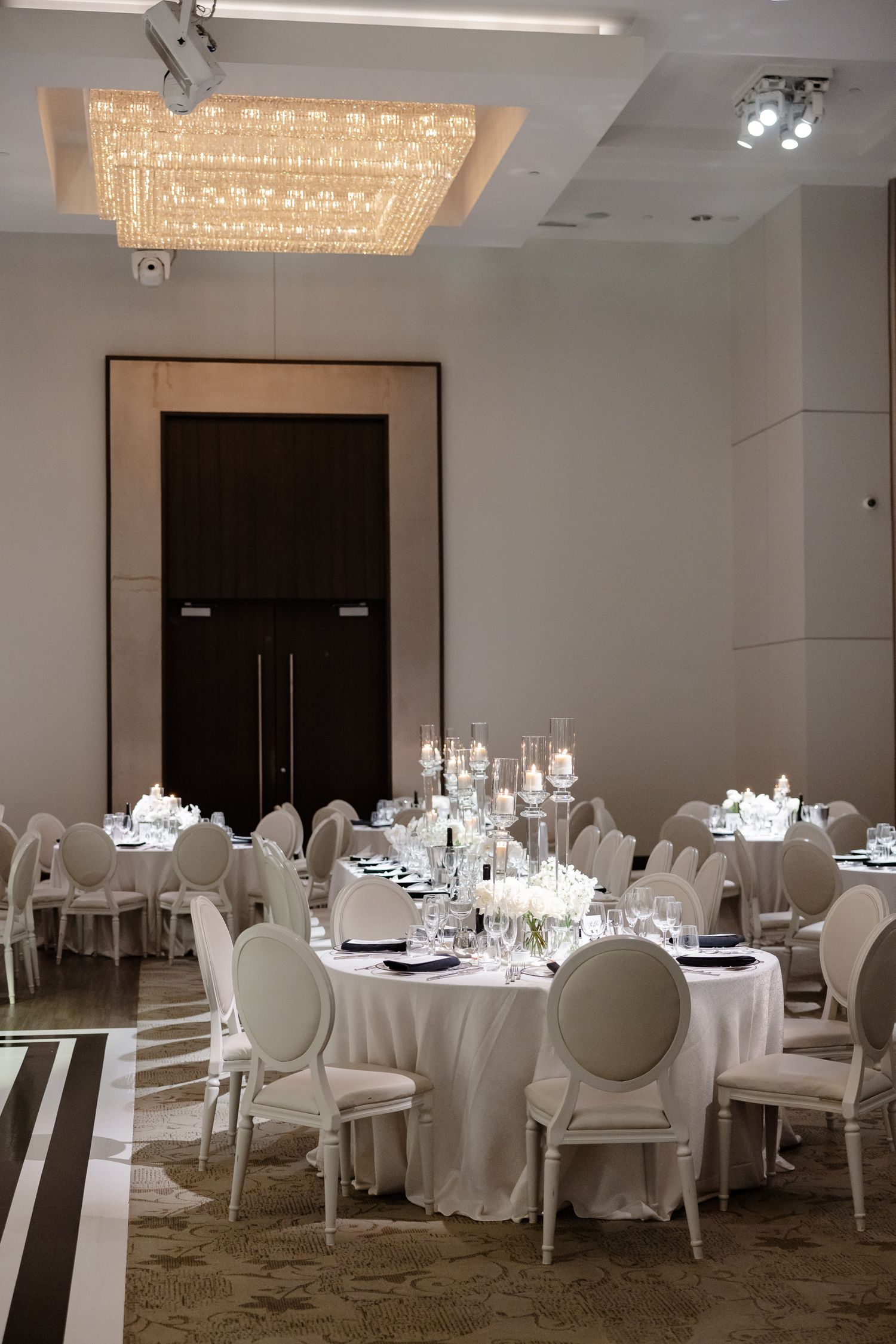 Elegant ballroom setup with round tables, white linens, Louis XVI chairs, and a crystal chandelier overhead.