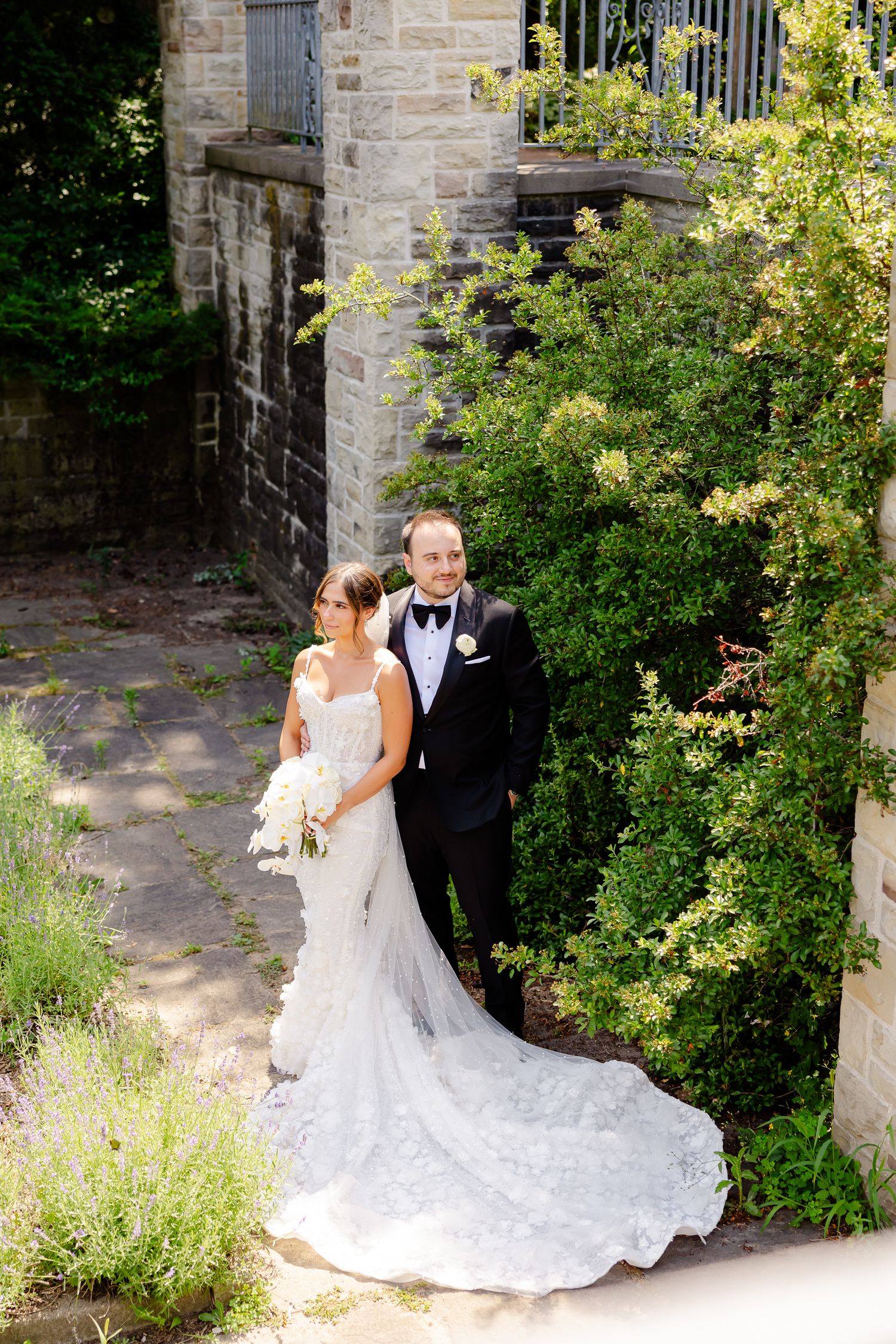 A newlywed couple poses in formal attire against a stone garden wall with lush greenery and trailing bridal gown train.