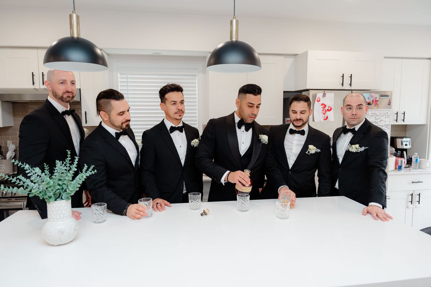 Group of groomsmen in black tuxedos gather around white kitchen counter.