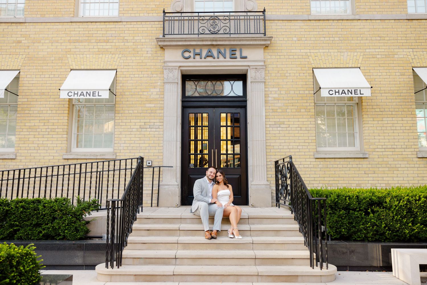 A sun-lit photo of a couple on the steps of a Chanel store with yellow brick facade.