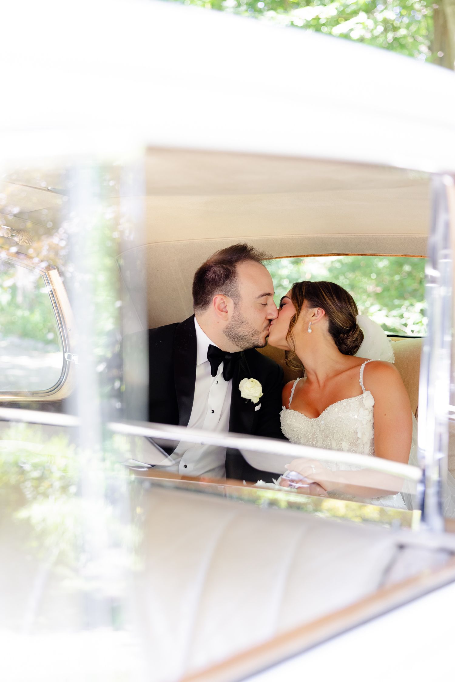 A romantic wedding couple shares a kiss in the back seat of a vintage car with a white boutonniere visible.