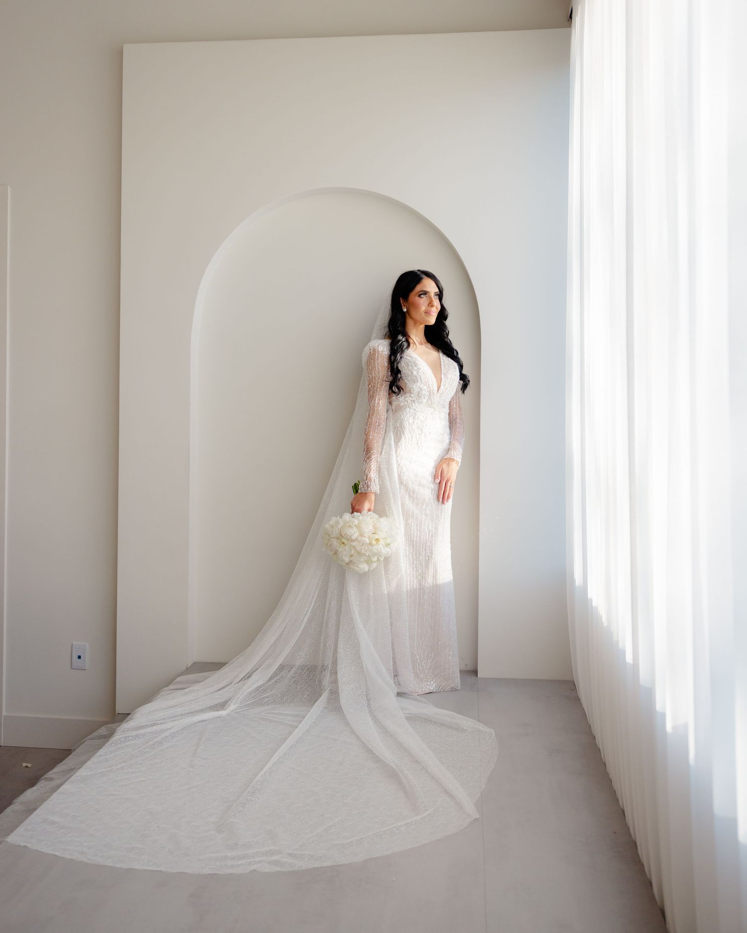 Bride in lace gown with cathedral veil standing in minimalist white archway.