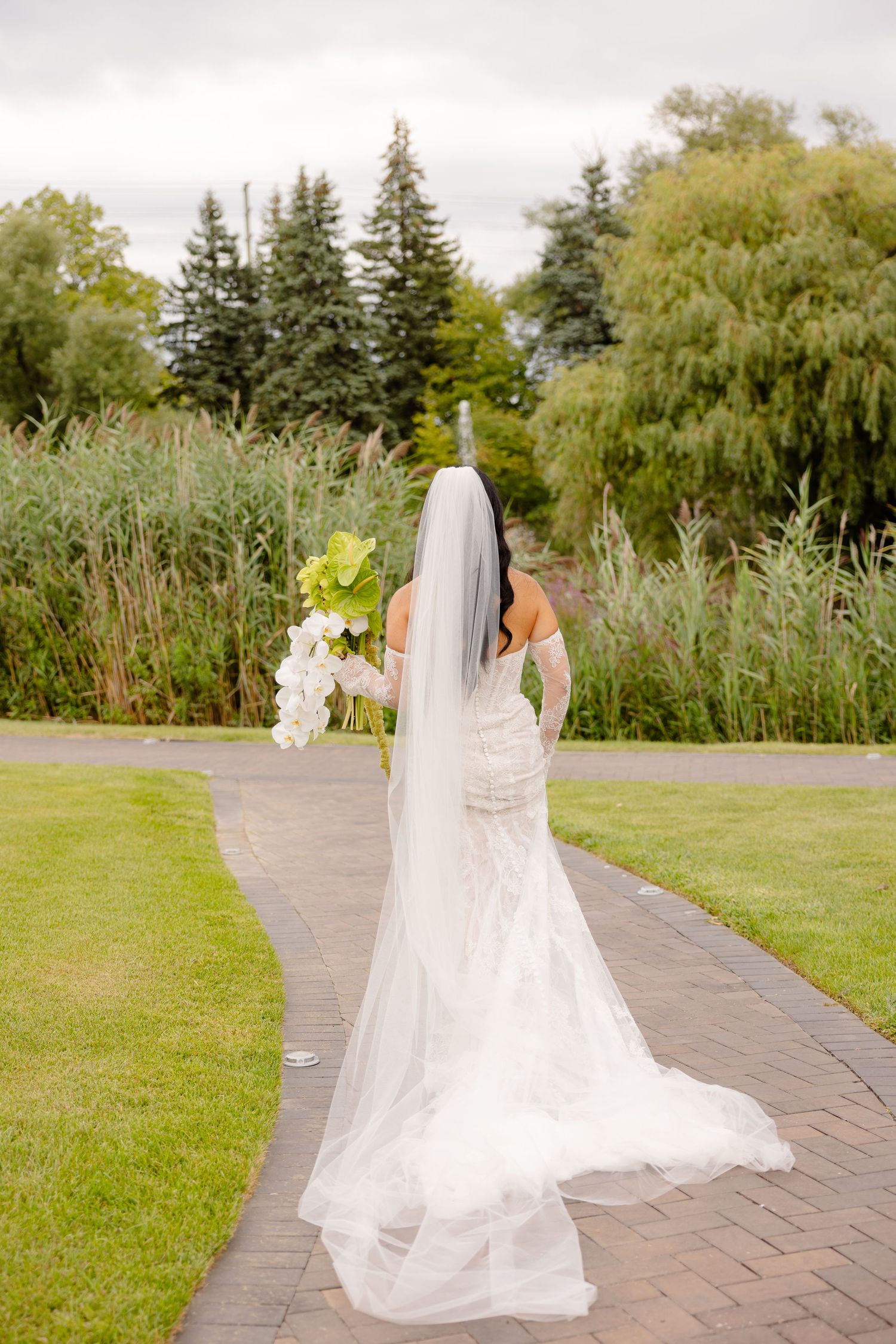 A bride in a flowing white wedding gown walks along a garden path with trees and greenery in the background.
