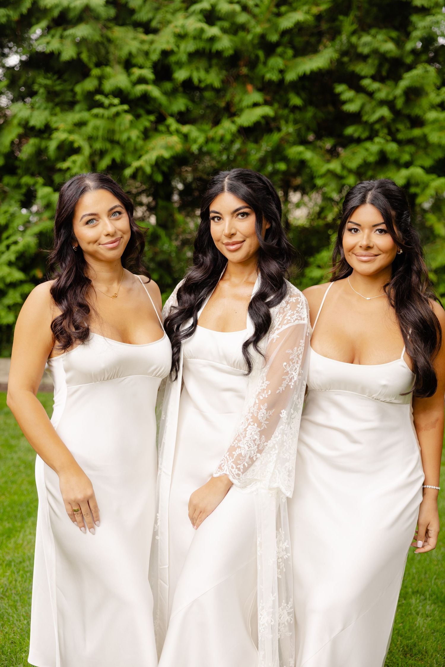 Three women in white dresses stand together smiling on a grassy lawn with green foliage in the background.