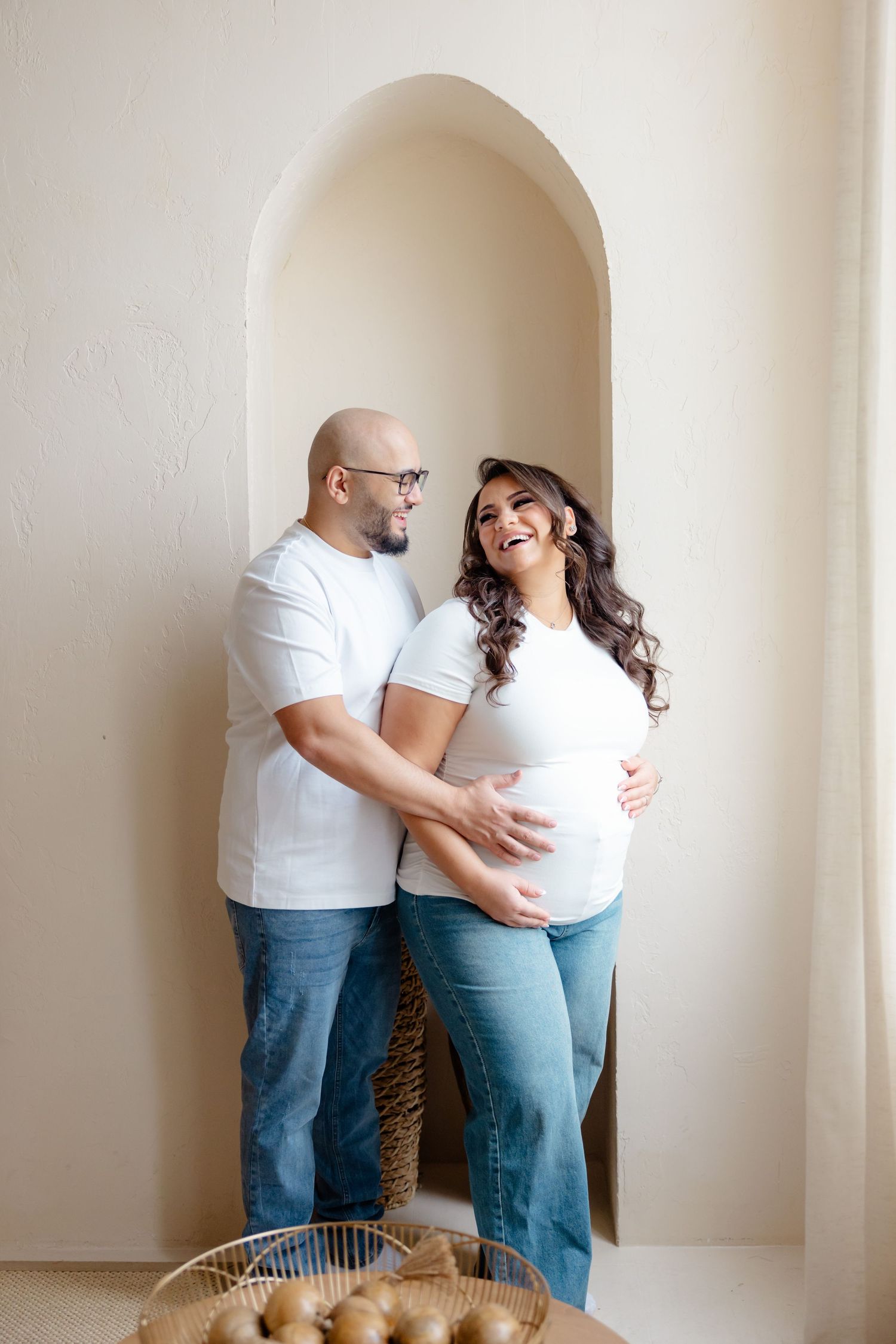 Expecting couple in white shirts and jeans share a joyful moment together in an arched alcove with a basket nearby.