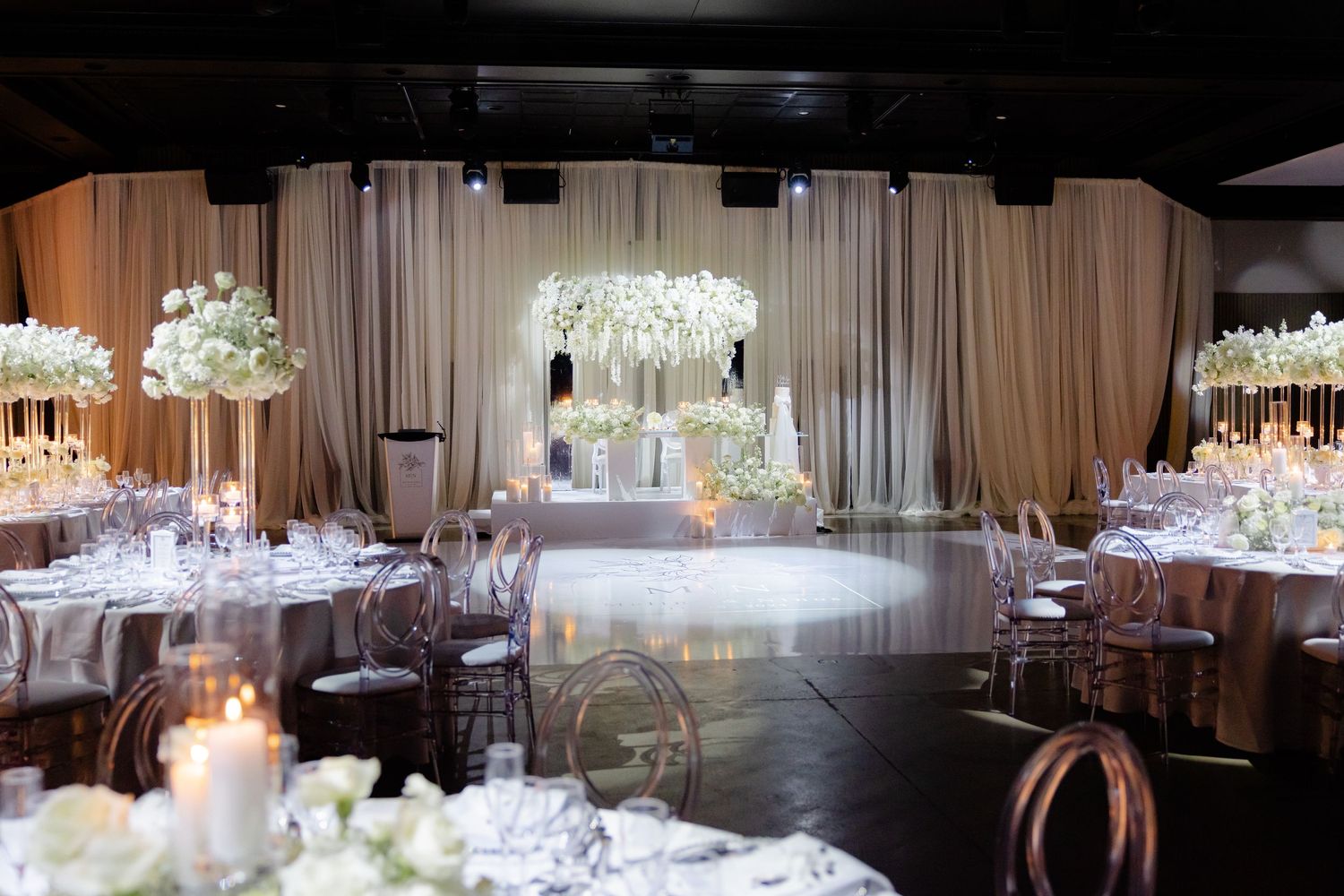 Wedding reception hall with draped fabric backdrop and crystal chandelier centerpiece.