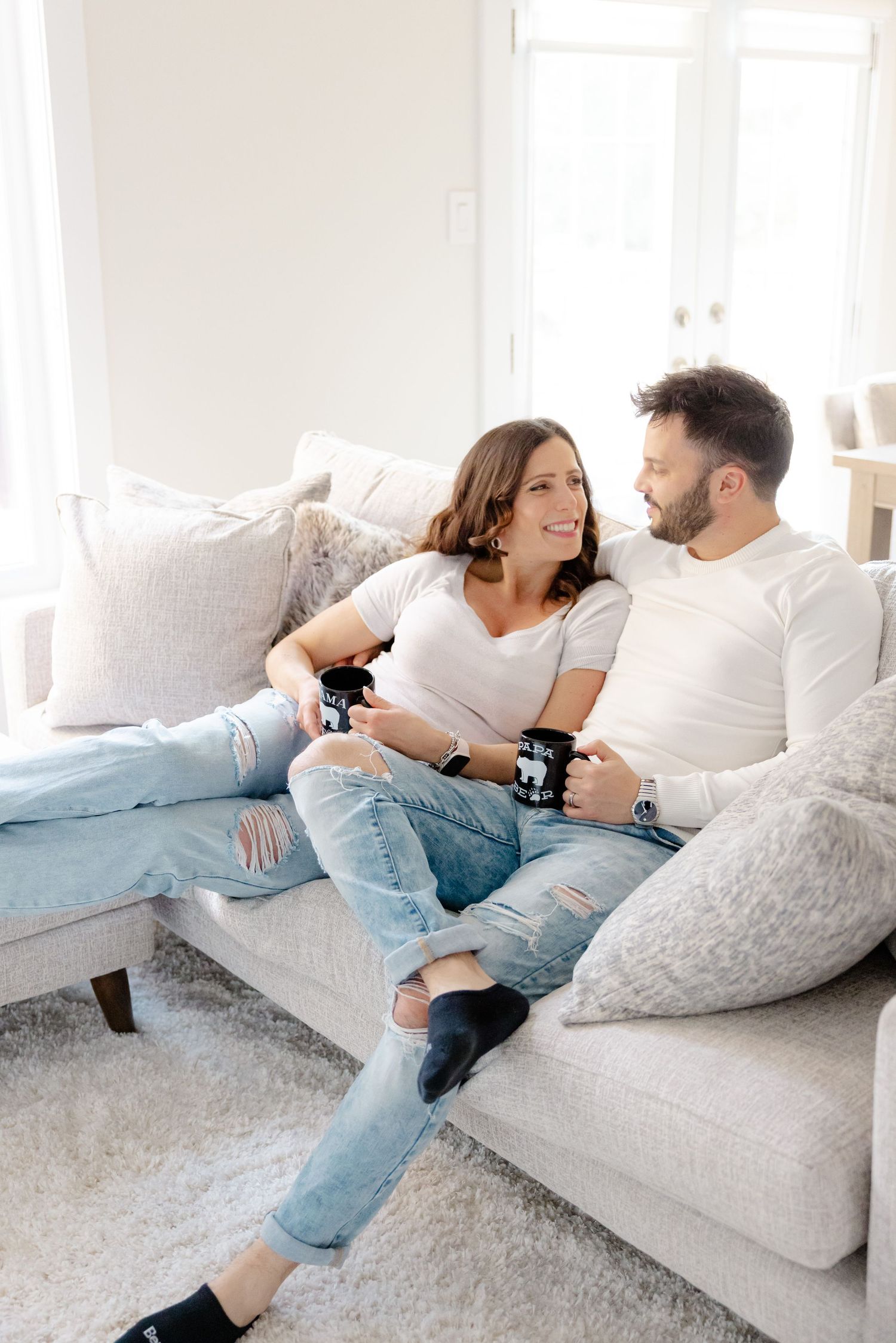 Couple relaxing on white couch in bright room, holding beverages and smiling at each other.