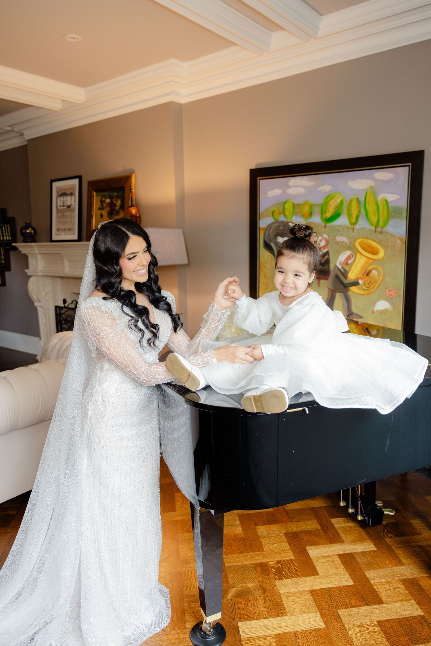 A bride helps a young attendant balance while sitting on a grand piano during wedding preparations.