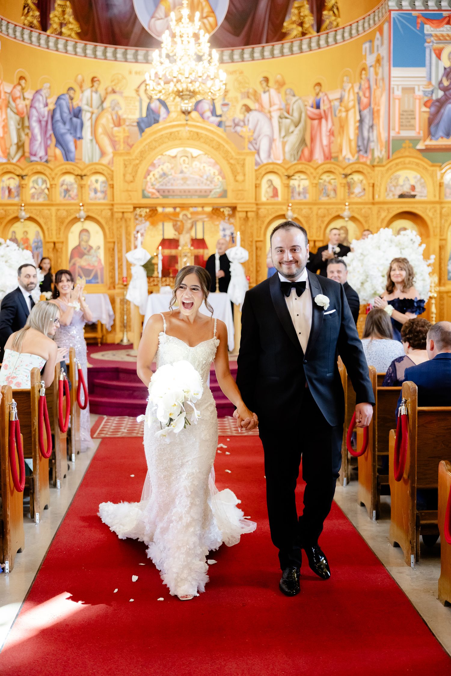 Newlywed couple walks down red-carpeted church aisle together surrounded by ornate gold iconography and a crystal chandelier.