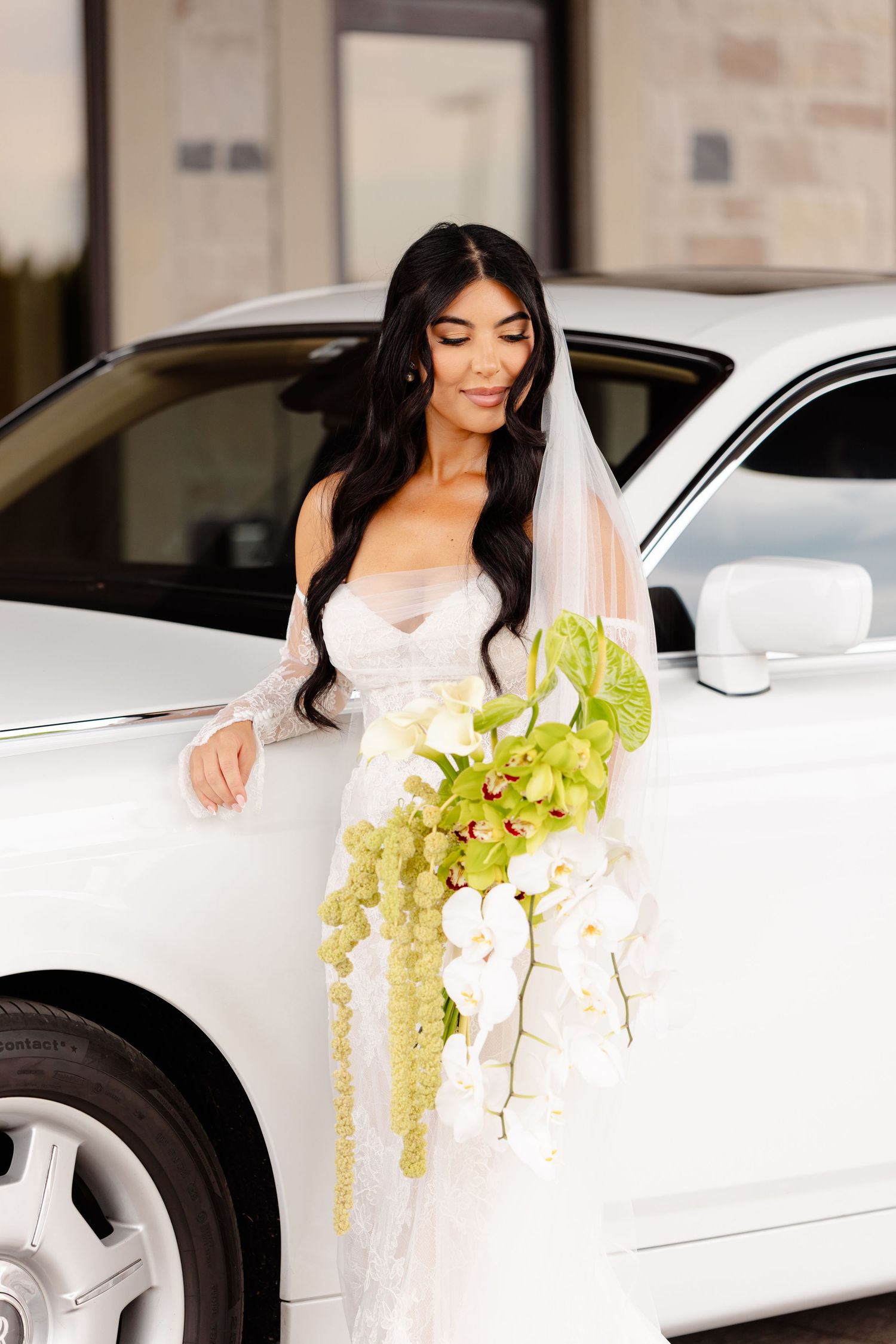 Bride in white dress poses with green floral bouquet next to white luxury sports car.