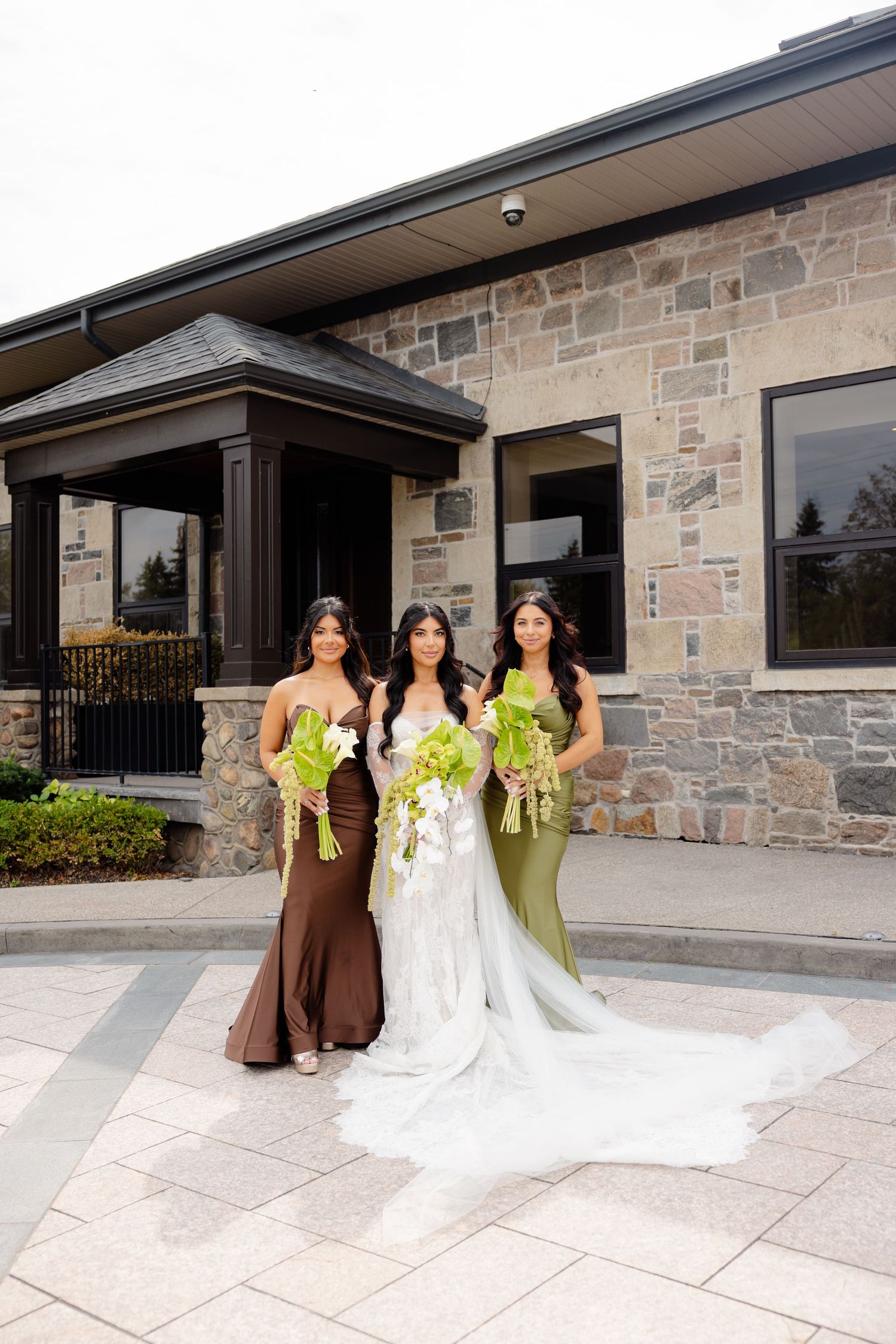 Bridesmaids in brown and sage green dresses stand with bride in white gown outside stone building during wedding.