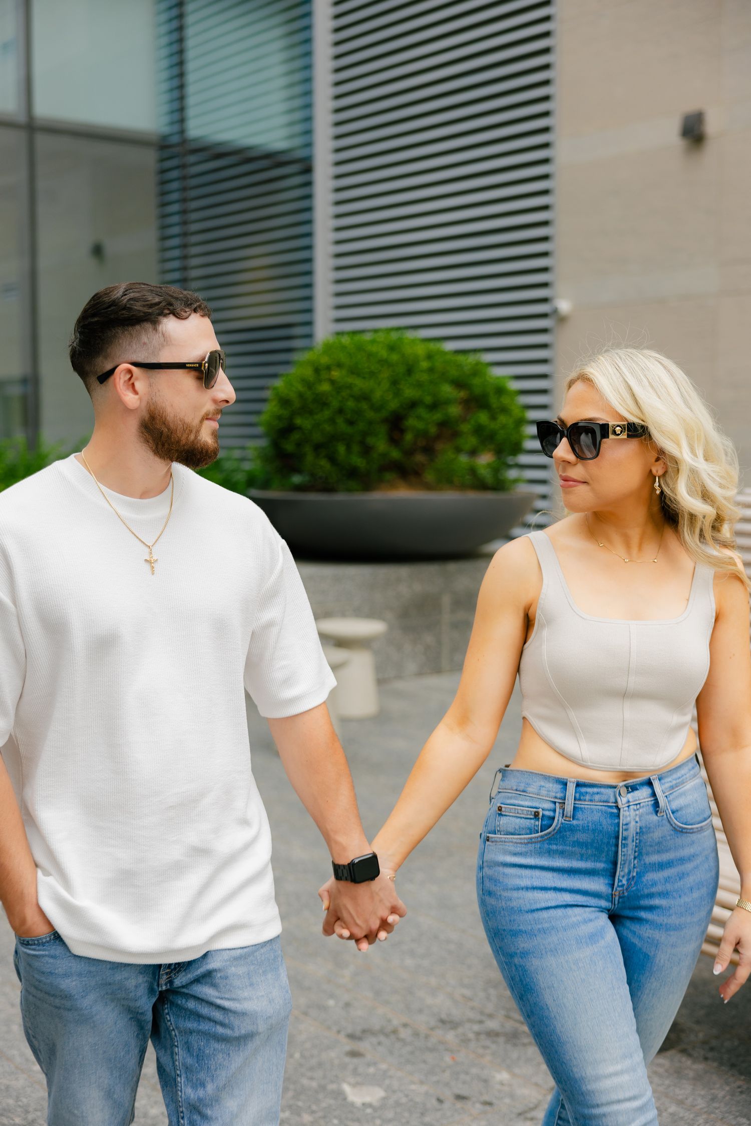 A couple holding hands and wearing sunglasses walk together outside a modern building.