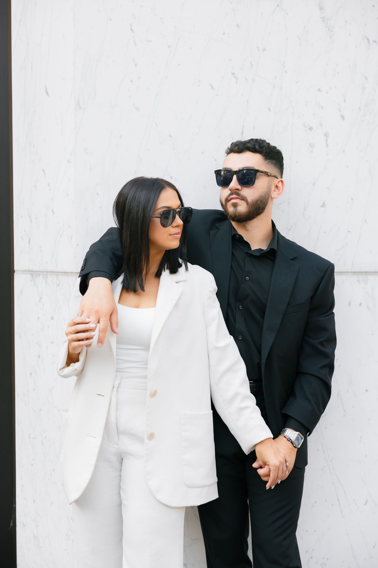 Couple in black and white outfits wearing sunglasses pose closely together against white wall.